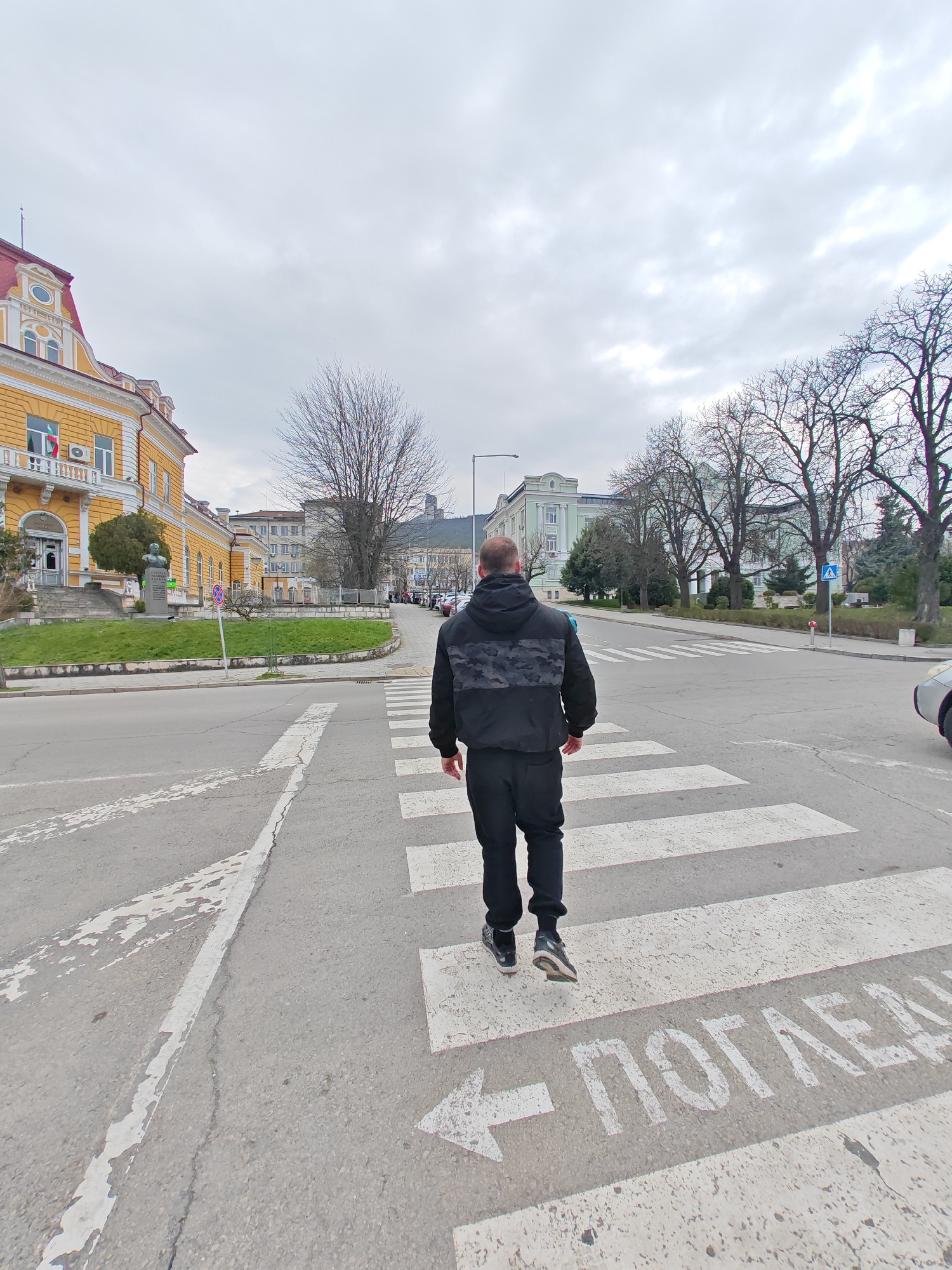 An adult male, viewed from the back, is observed walking across a pedestrian zebra crossing on an asphalt street in Shumen, Bulgaria, under an overcast sky. He is wearing a black hooded jacket with a grey camouflage pattern on the upper back, black athletic trousers, and dark athletic shoes. His right foot is slightly elevated in mid-stride. White painted markings on the asphalt include the zebra stripes, a white arrow, and the Cyrillic word "ПОГЛЕДИ" (Look/Look around). The street markings show signs of wear.
To the left of the pedestrian, a multi-story yellow building with white architectural details and a red tiled roof, identifiable as the Shumen Regional Administration building, dominates the background. A Bulgarian flag is affixed to a second-story balcony on this building. In front of the building, a dark-colored bust on a pedestal is situated within a grassy median, alongside a blue circular 'no stopping' sign. Bare deciduous trees are visible behind and to the left of the yellow building.
To the right, a street extends into the distance, lined by numerous bare deciduous trees. Further down the street, a pale green public building with a flat roof is visible. A blue triangular pedestrian crossing sign is posted on the far side of this street. A partial view of a light-colored vehicle (silver/gray) is visible on the far right edge of the frame. Hills are discernible in the far distance beneath the uniformly cloudy sky.