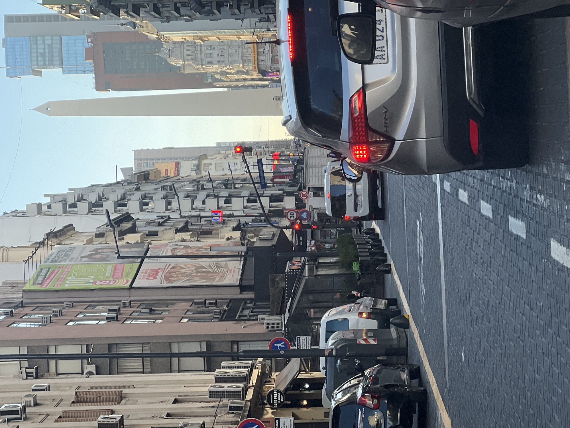 The image captures a street scene in Buenos Aires, Argentina. In the distance, the Obelisco, a historical monument, rises prominently against the skyline. The foreground is dominated by traffic, featuring a silver Honda HR-V with license plate AA UZ4, a white service vehicle with license plate AF 871 TO, and a blue car. The street is paved with a dark gray brick pattern. Pedestrian signs and a "30" speed limit sign are visible along the street. Buildings line both sides of the road, with billboards and advertisements on top. Various details such as air conditioning units, shop signs (QURO PRETO "EL MEJOR CAFE") and traffic lights add to the urban atmosphere.