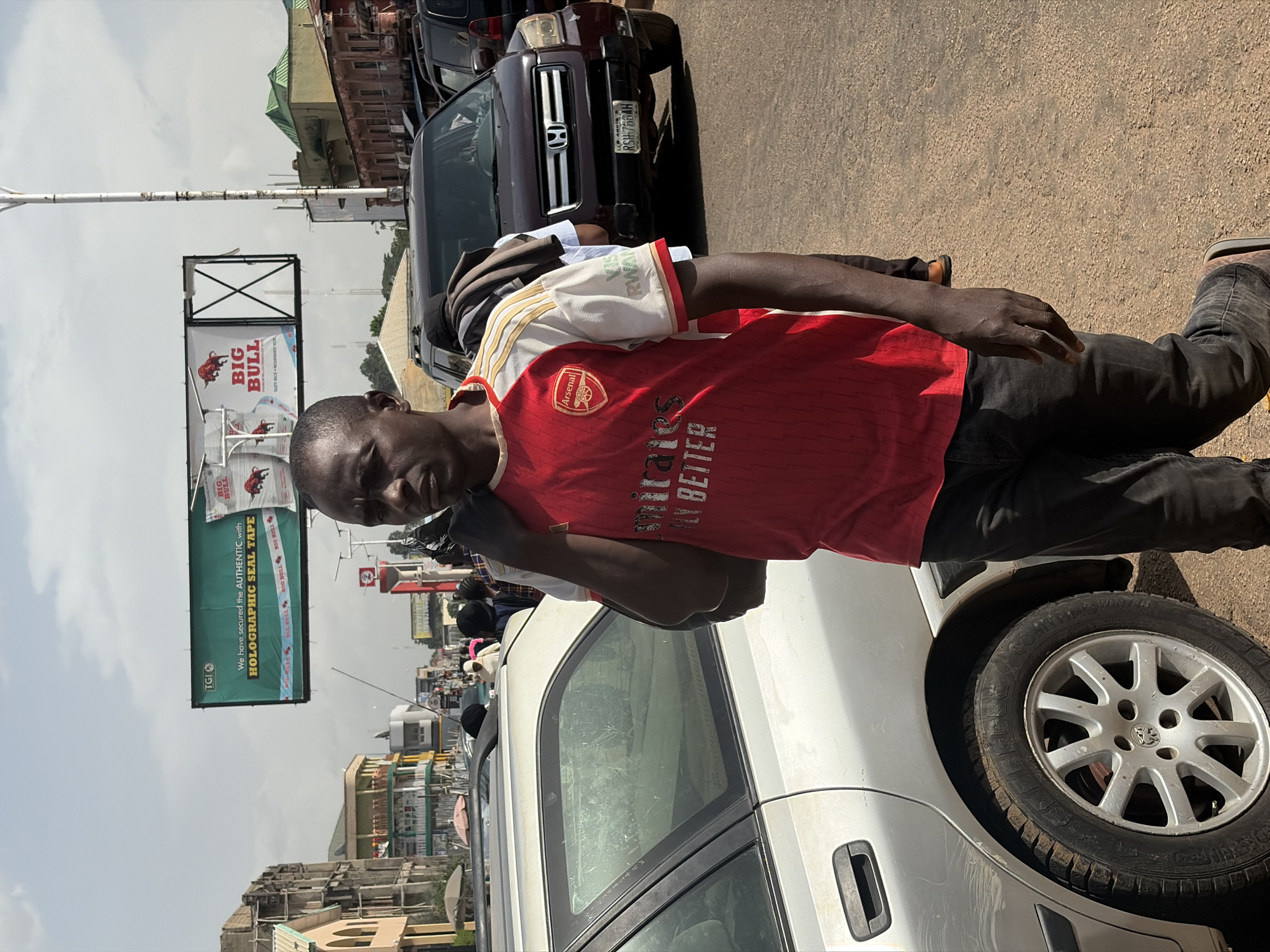 The media file captures a vertical outdoor scene on a street in Jos, Nigeria. The main subject is a young Black man wearing a red and white Arsenal football jersey and black jeans. He is standing near a white car and appears to be on his phone, with his hand raised to his ear. Behind him, a billboard advertises "BIG BULL" with images of bulls and text about "TASTY RICE" and "HOLOGRAPHIC SEAL TAPE". Other vehicles, including a dark SUV with the license plate "RSH-786AH", are visible in the background, suggesting a busy street. Buildings with distinctive architecture, some appearing older, line the street. The sky is overcast, indicating a cloudy day. The scene appears to be during daylight hours. The overall atmosphere seems to be that of an everyday street scene in an urban environment in Nigeria.