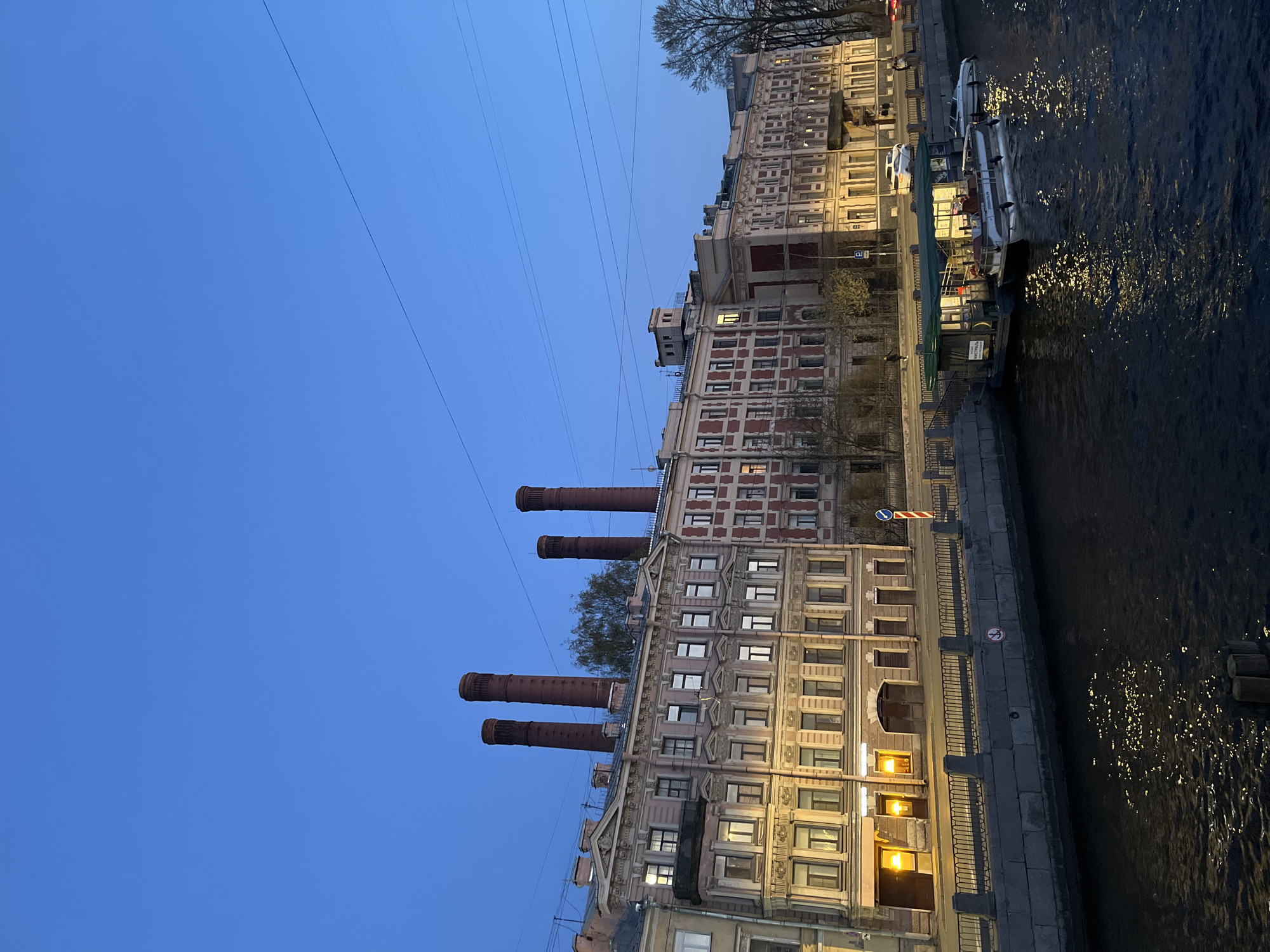The image depicts an urban waterside scene at dusk or early evening, characterized by a deep blue sky. A row of multi-story buildings lines the left bank of a dark canal or river, their windows illuminated by artificial light, casting warm glows. The architecture varies, featuring reddish-brown brickwork, light-colored facades, and decorative elements.
A prominent building in the middle of the frame is topped with five tall, reddish-brown brick chimney-like structures, suggesting a former industrial purpose. Along the right bank of the waterway, several boats are docked, including a larger passenger vessel with a green canopy. The dark water shimmers with numerous reflections of the building lights.
Multiple thin power lines stretch across the clear sky. Bare trees are visible between and behind some of the buildings. Visible signs on the embankment include a blue circular sign with a red diagonal bar, a "No Swimming" sign (a person in water with a red line through it), and a blue parking sign with a white 'P'. Text on the side of one boat appears to include "ОЧНЫЕ КОМПЛЕКСЫ" and possibly "ПРОГУЛОЧНЫЕ", indicating excursion or pleasure boats. The overall atmosphere is tranquil.