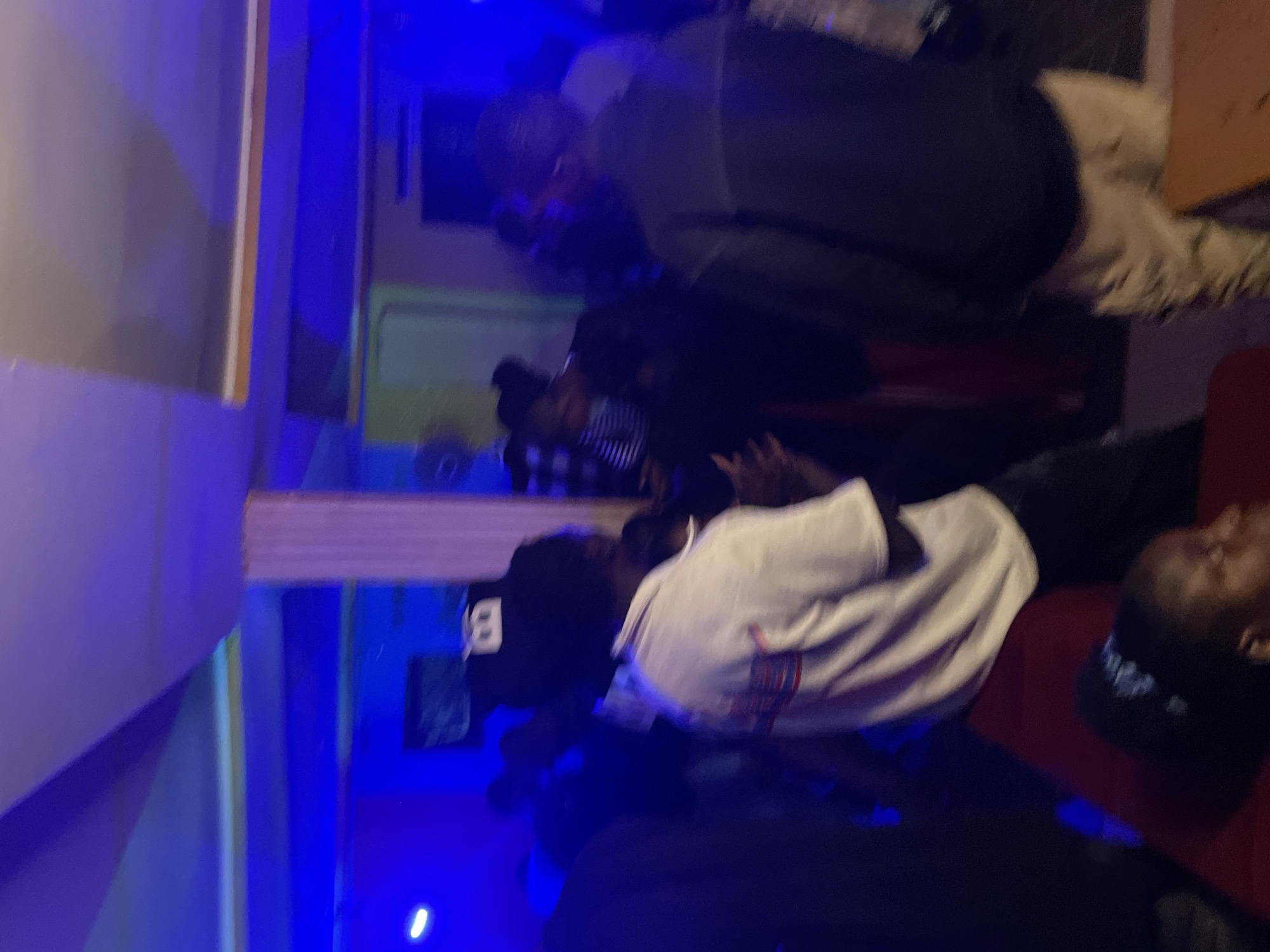 The image depicts an indoor scene, possibly a lounge or club, in Durumi, Nigeria. The environment is dimly lit, with prominent blue and purple lighting casting an atmospheric glow. Several individuals are present, with their features somewhat obscured by the low light and blur. In the foreground, a person is seen from the back, wearing a white t-shirt with red and blue stripes on the sleeve and black pants. To their right, another individual is visible from the back, wearing a dark jacket and light-colored pants. Other people are scattered throughout the background, some appearing to be standing and socializing. A red seating area is visible in the lower left portion of the frame. The overall impression is of a casual gathering or social event.
