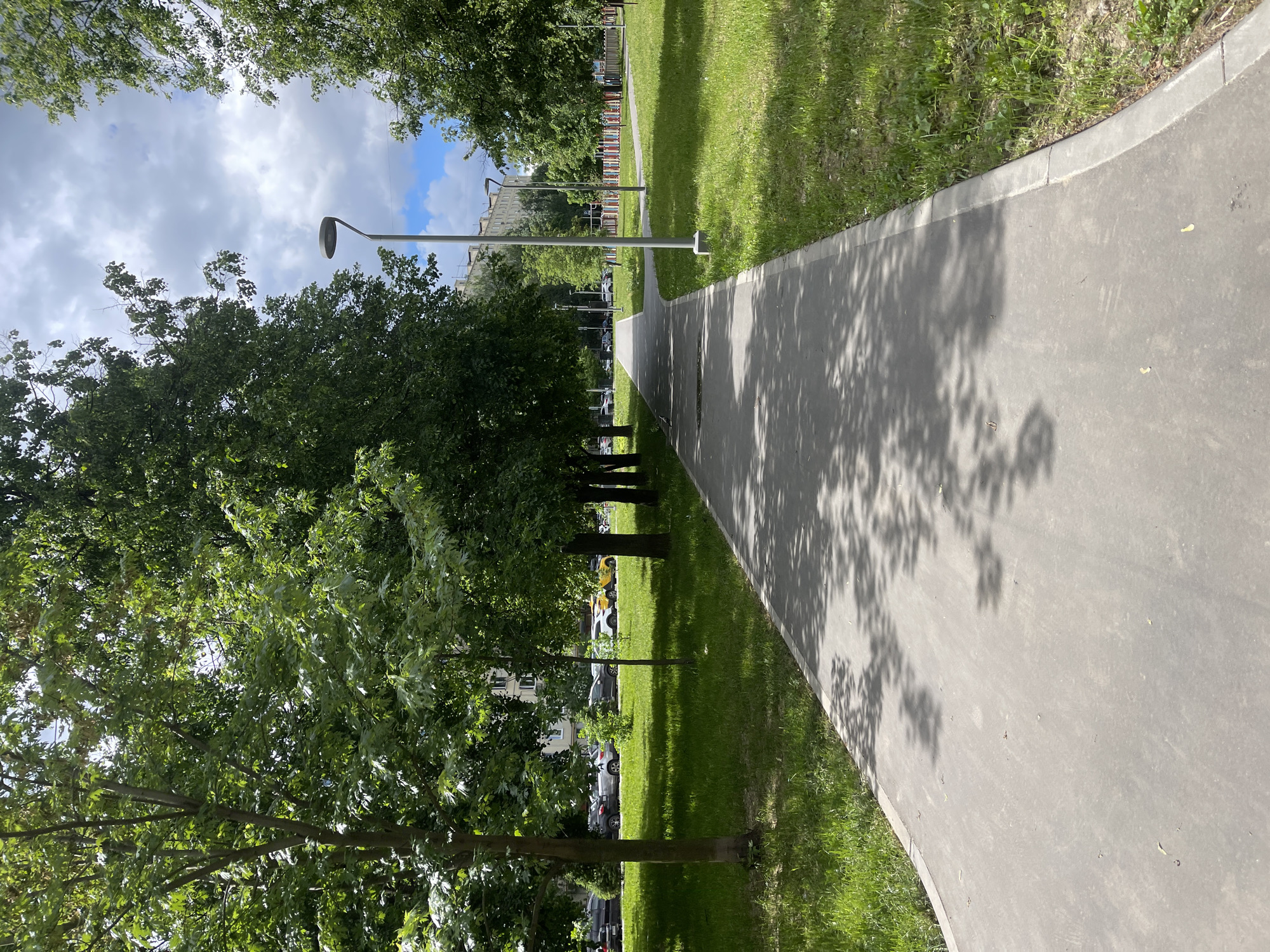 A paved pathway bisects a grassy lawn in an urban setting. The path curves slightly to the right, and tree shadows are visible upon its surface. Trees line the left side of the path, obscuring buildings and parked cars. Lampposts run parallel to the path on the left. The sky above is partly cloudy. The location is Moscow, Russia.