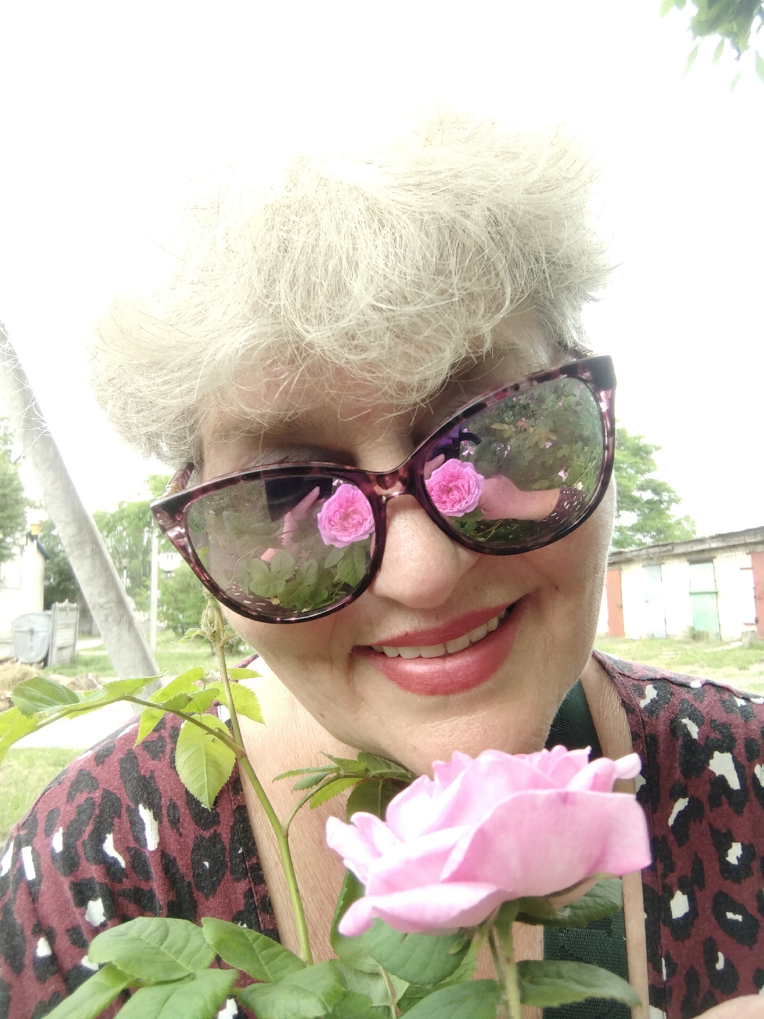 A close-up, high-angle selfie captures a smiling older woman outdoors in Horishni Plavni, Ukraine, on a bright day. She has short, voluminous, light grey or white hair and is wearing purplish-framed sunglasses and a maroon and black patterned top, possibly a leopard print. Her lips are painted with red lipstick, and she displays a cheerful, open-mouthed smile.
In her right hand, she holds a delicate light pink rose with green leaves, positioned prominently in the lower right foreground. Notably, the lenses of her sunglasses reflect the scene in front of her, clearly showing two vibrant pink roses and lush green foliage, along with what appears to be her hand holding a rose.
The background is somewhat overexposed but reveals a rustic or semi-rural setting. Visible elements include a utility pole on the left, hints of green trees and grass, and several single-story buildings in the distance, resembling garages or sheds with various colored doors (e.g., green, red). The bright lighting suggests it is daytime. The woman appears happy and content. No discernible text is visible in the image.