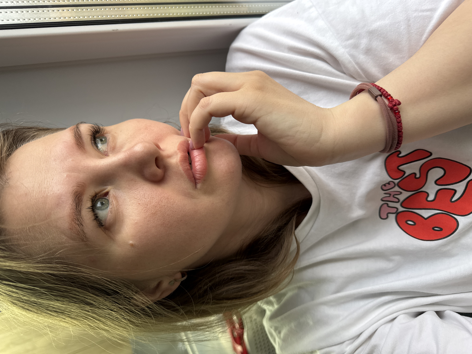 A young woman with fair skin and blonde hair is depicted in a close-up, slightly angled shot. Her eyes are light blue, and she is touching her lips with her fingers. She is wearing a white t-shirt with a red and pink graphic design. On her wrist, she has two bracelets, one a maroon elastic band and the other a braided string with red beads. The background is blurred, but a window frame is visible. The location is not discernible from the image alone.