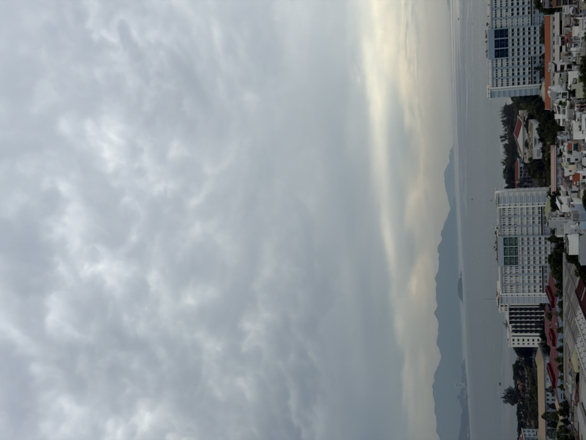 The image captures an overcast day in Nha Trang, Vietnam, looking out over the city towards the sea and a mountainous coastline. The foreground is dominated by the rooftops of urban buildings, characterized by a mix of white structures with visible window grids, some with orange or red roofs, and larger, multi-story white and blue buildings. Amidst the urban sprawl, there are glimpses of green trees and foliage.
In the middle ground, the calm, grey sea stretches towards the horizon. Along the coast, a series of dark, hazy mountains forms a scenic backdrop. A small, dark boat can be observed on the water. The sky is filled with heavy, grey clouds, creating a diffused light that softens the overall scene. The atmosphere suggests a potentially humid or rainy day, typical of Nha Trang's tropical climate. No people are visible in the image.