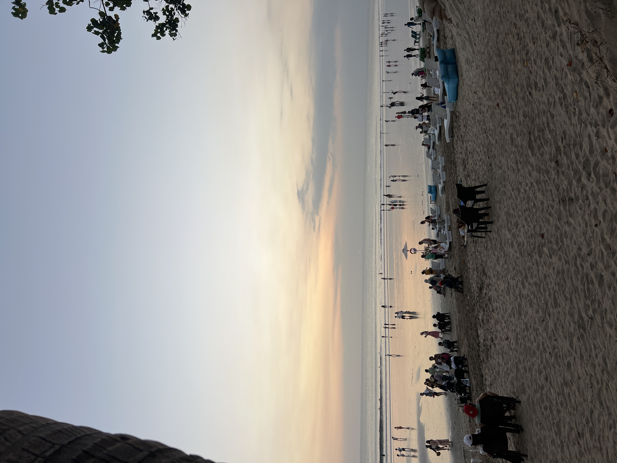 A wide-angle, eye-level shot captures a beach scene at sunset in Kuta, Indonesia. The sky is a gradient of soft blues and pale oranges, with a few wispy clouds near the horizon. The ocean is calm with gentle waves breaking on the shore. A wet, reflective sandbar stretches across the middle ground, populated by dozens of silhouetted figures walking or standing. In the foreground, the sand is drier and darker, with a scattering of chairs, tables, and lounge furniture. Several groups of people are seated on the sand, some conversing, others relaxing. A few individuals are standing near the water's edge. The overall atmosphere is serene and communal.