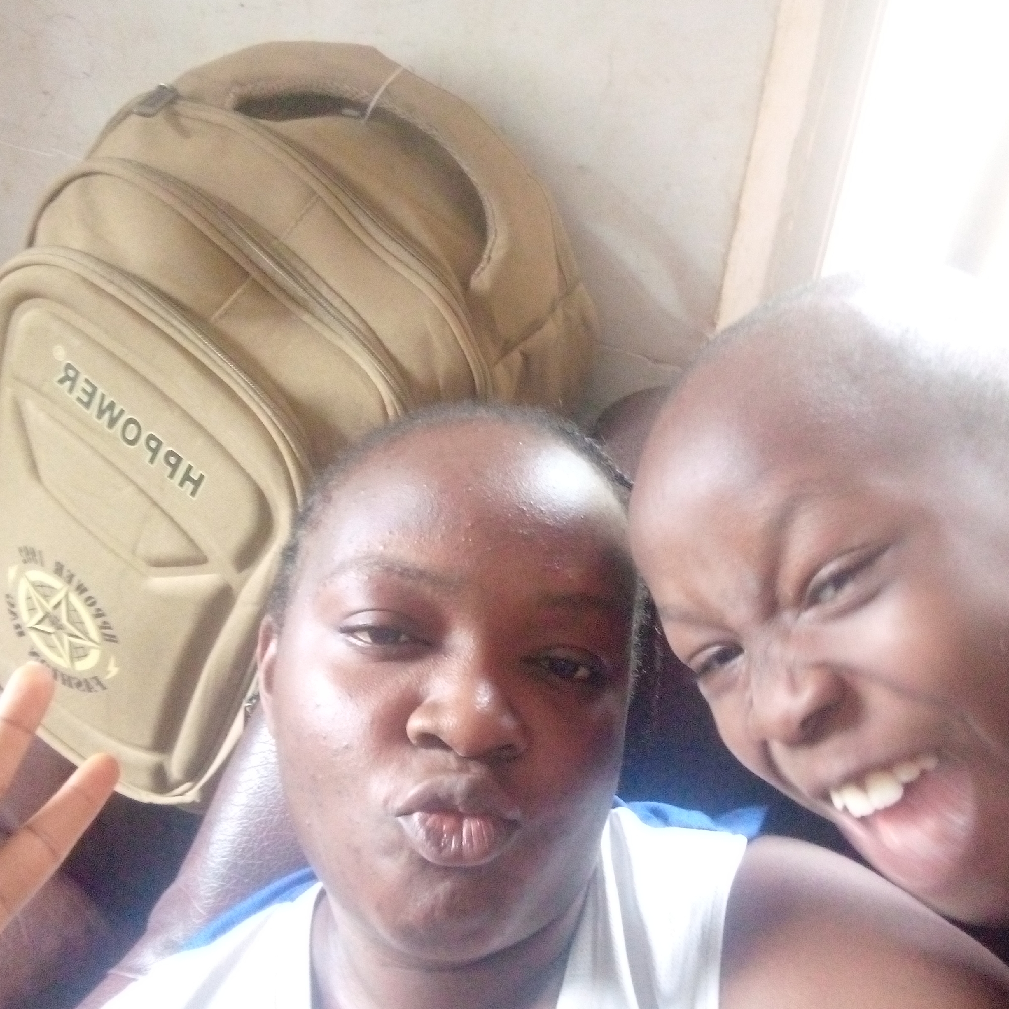This is a close-up, high-angle selfie featuring two individuals and a backpack, creating a lighthearted atmosphere.
The primary subjects are an adult and a child, both with dark complexions. The adult is in the foreground on the left, looking towards the camera with pursed lips, as if making a "kiss" face. They are wearing a white top, and their left hand is partially visible, showing a peace sign. To the right and slightly behind the adult is the child, making a playful, wide-mouthed grimace, showing their teeth and conveying a sense of joy or silliness.
Behind the individuals, a light brown or khaki-colored backpack is prominently visible. It features several zippered compartments and shoulder straps. On the backpack, mirrored text is visible. When unmirrored, the main text on the top section appears to read "HBPOWER". Below this, there's a circular logo resembling a compass rose, which also contains text. The unmirrored text within and around the logo includes "HBPOWER", "1984", and partially decipherable letters that suggest "REVERB". The background behind the backpack is a light-colored wall.
The lighting appears bright, suggesting daytime, possibly from an indoor light source or a nearby window. The overall mood of the image is cheerful and informal, capturing a candid, playful moment between the two individuals.