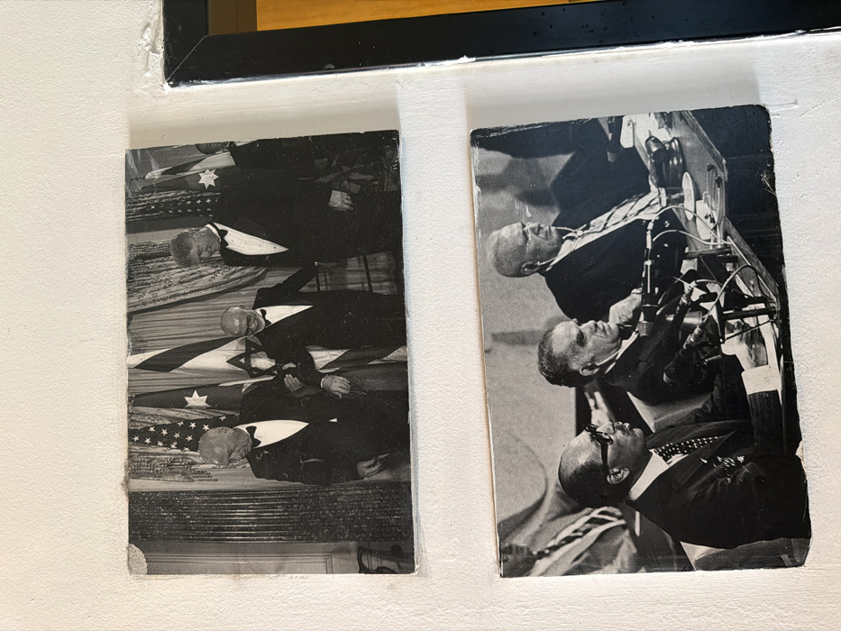 Two aged, black and white photographs are mounted vertically on a white, textured wall in a location within Tel-Aviv, Israel.
The upper photograph depicts three male figures, identified as Shimon Peres (left), Yitzhak Rabin (center), and Bill Clinton (right), all wearing tuxedos with bow ties. Rabin is being supported on his right arm by Peres. All three individuals are smiling and looking forward. In the background, draped curtains and several national flags are visible, including the flag of the United States and the flag of Israel. This scene likely captures a moment during the signing of the Oslo Accords or a related diplomatic event at the White House. The photograph exhibits wear, with curled edges and minor surface damage.
The lower photograph features three prominent male figures, identified as Anwar Sadat (left), Menachem Begin (center), and Jimmy Carter (right), seated at a long table. All are wearing suits and ties. Sadat wears dark sunglasses. The table is equipped with multiple microphones, papers, and a wooden gavel is visible on the right side. The scene is consistent with the Camp David Accords discussions or signing ceremony. This photograph also shows signs of age, with slightly curled edges and minor surface imperfections.
A partial view of a black-framed object, likely another picture, is visible on the right edge of the overall image, adjacent to the lower photograph.