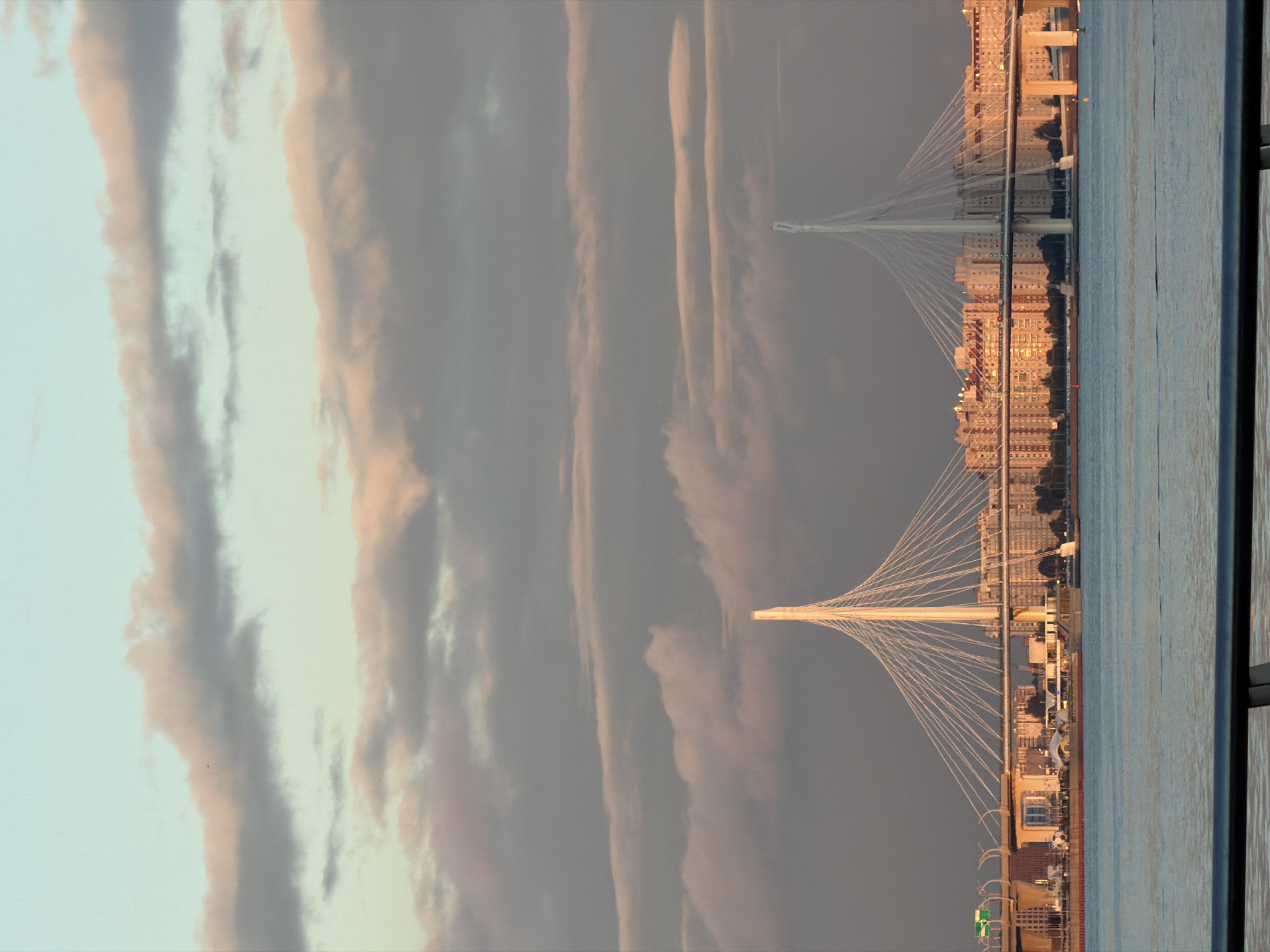 The image captures a wide shot of a cable-stayed bridge in Saint Petersburg, Russia, spanning across a body of water. The bridge features two prominent white towers with cables fanning out to support the roadway. In the background, a dense cluster of residential buildings with light-colored facades and red-tiled roofs is visible. The sky is filled with a mix of soft, pastel-colored clouds, suggesting either sunrise or sunset. The water in the foreground is calm and reflects the sky and the bridge. A dark railing runs along the bottom of the frame, indicating the vantage point is from a balcony or a similar elevated structure. There are no people or animals visible in the frame.