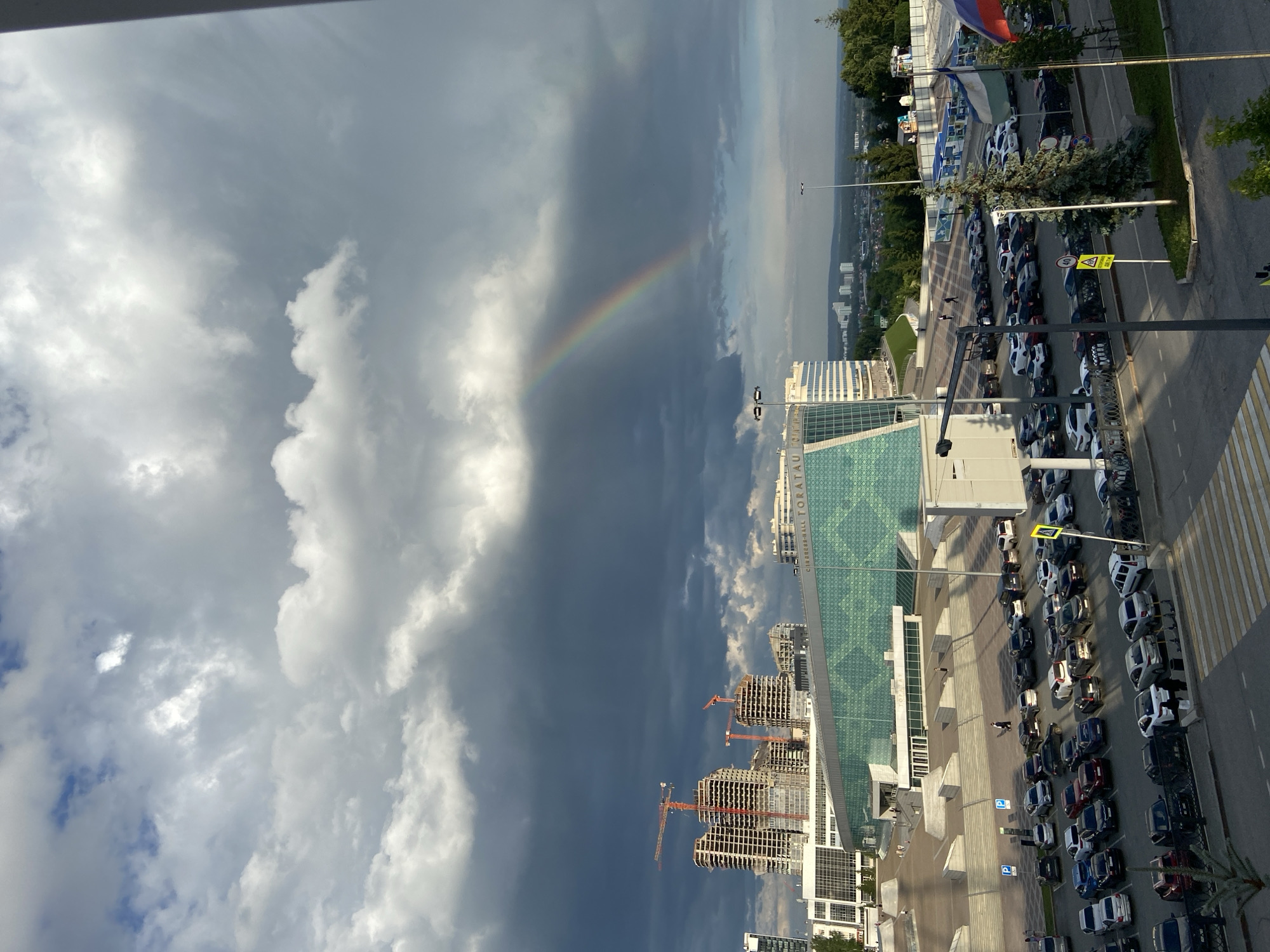 The image depicts a panoramic view of the area surrounding the Toratau Congress Hall in Ufa, Russia. In the foreground, a parking lot is filled with numerous vehicles. Beyond the parking lot, the Toratau Congress Hall stands, a modern building with a distinctive glass facade. Construction cranes are visible in the background, indicating ongoing development in the city. A rainbow arches across the sky, suggesting recent rainfall. The overall scene captures an urban landscape under a dramatic sky.