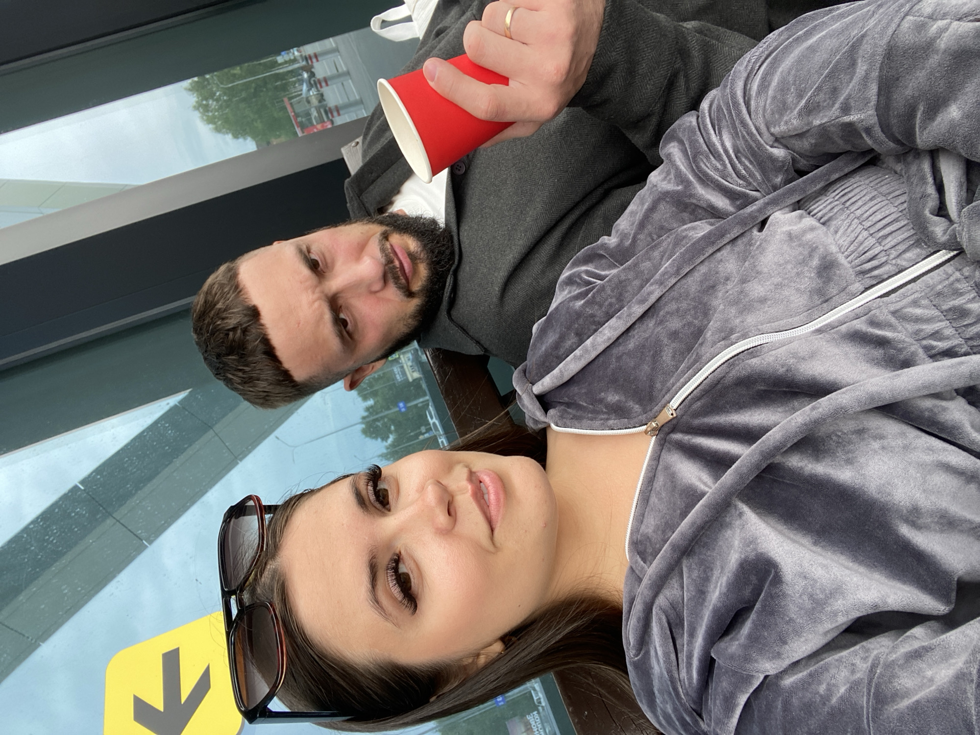 A young woman with long dark hair and false eyelashes is taking a selfie with a man. She is wearing a grey velour tracksuit, and sunglasses are perched on top of her head. The man, who has a beard and short dark hair, is wearing a dark jacket over a white t-shirt and is holding a red disposable cup. They are seated on a bench, with a large window behind them showing an outdoor scene. A yellow directional arrow sign with a black arrow pointing left is visible on the window. The background outside the window shows trees, a street with cars, and some poles. The setting appears to be a public waiting area, possibly an airport or train station. There is no information or visual cue that indicates the location as Новое Мартьяново, Russia.