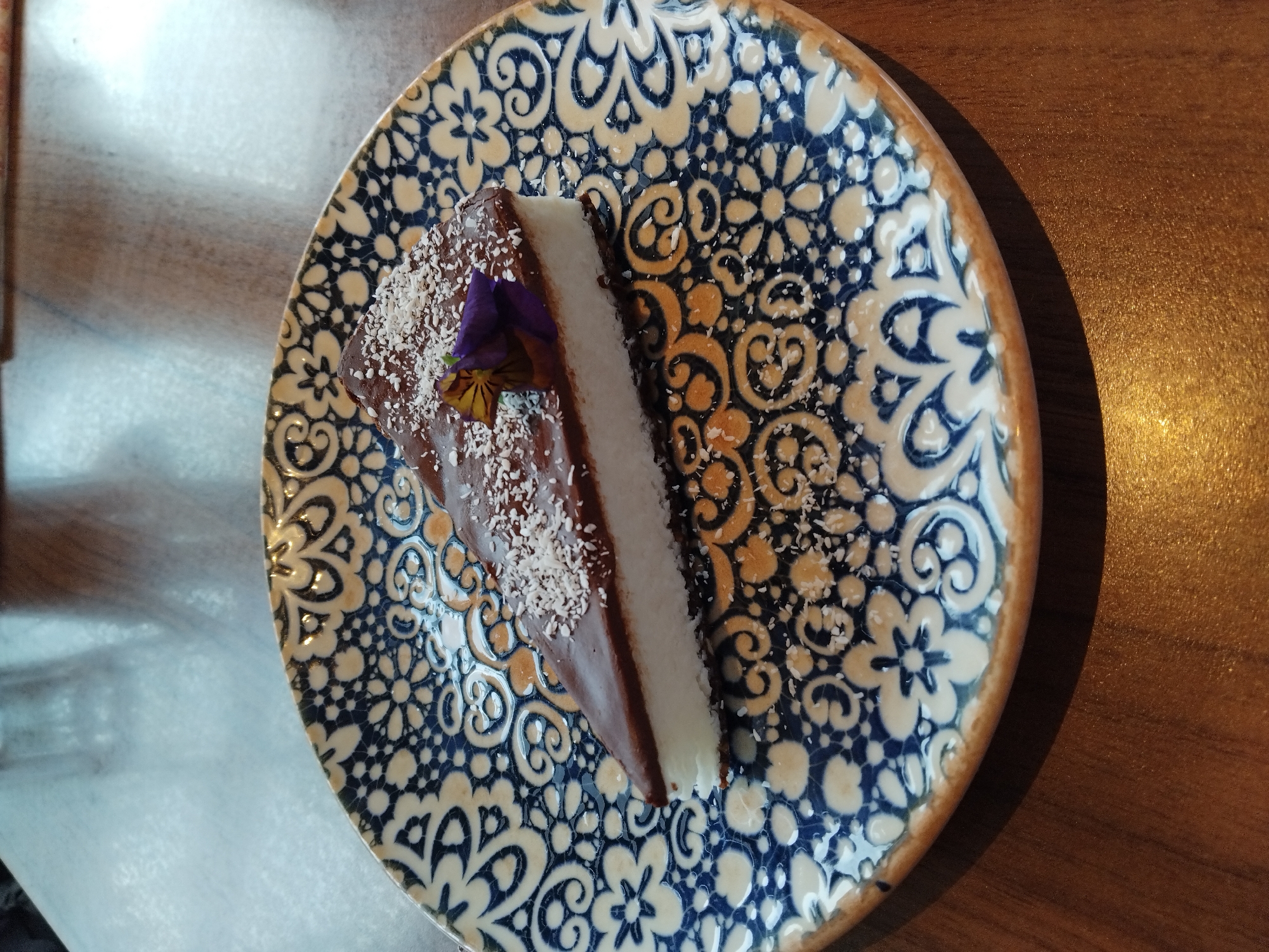 A top-down shot showcases a slice of cake on a patterned ceramic plate. The cake has a dark chocolate top, a white creamy layer, and is sprinkled with shredded coconut. A small purple and yellow flower garnishes the cake. The plate has a blue and white floral pattern with an orange-brown rim. The plate rests on a wooden surface.
The scene is inferred to be in Varna, Bulgaria, but there is no conclusive evidence for this location in the image.