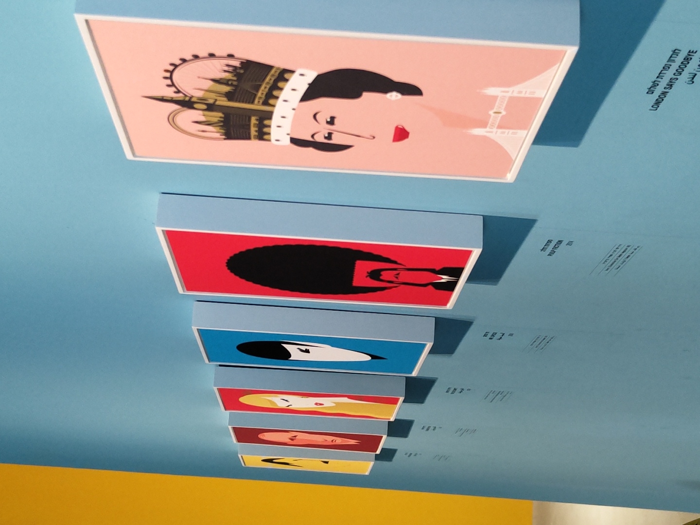 The image shows a row of framed artwork displayed on a light blue wall, with a bright yellow wall to the left. The artworks are stylized portraits with bold colors and minimalist designs. From left to right, the first artwork is yellow with a silhouette of a face. The next few are red, yellow, and blue, featuring abstract representations of human heads with distinct hairstyles. The fourth artwork in the row depicts a person with a large black afro. The largest and most prominent artwork on the right is a pink portrait of a woman wearing a crown that incorporates iconic London landmarks like Big Ben and the London Eye. She is also wearing a pearl earring and a necklace. Below the artwork, there are labels in Hebrew and English. One label clearly reads "PULP FICTION 2014". Another reads "LONDON SAYS GOODBYE". The context suggests this is an exhibition in Tel Aviv, Israel, likely showcasing pop culture-inspired art. The lighting is even, highlighting the artwork and the wall.