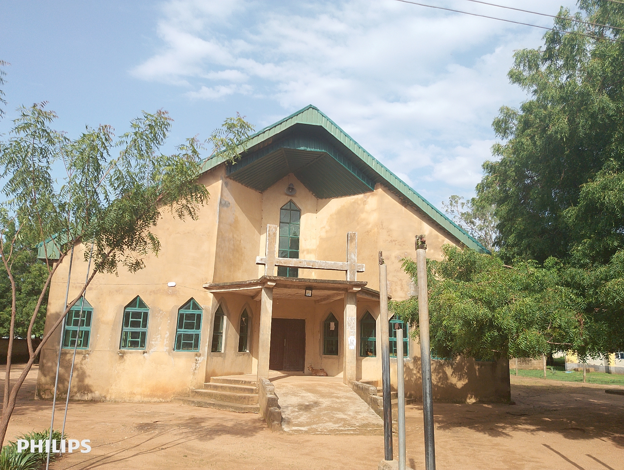 A wide, eye-level, outdoor shot captures a tan, stucco building with a green, corrugated metal roof. The building appears to be a church, with a prominent steeple-like structure at the front. Multiple arched windows with green frames punctuate the facade. A set of concrete stairs leads up to a covered entryway with double wooden doors. The ground in front of the building is a dusty, light brown earth, with several leafless trees casting shadows. A large, leafy green tree stands to the right of the building, its branches extending over the roofline. The sky above is a clear blue with scattered white clouds. In the lower left corner of the image, the word "PHILIPS" is visible in white text. The scene appears to be in a rural or semi-rural setting.