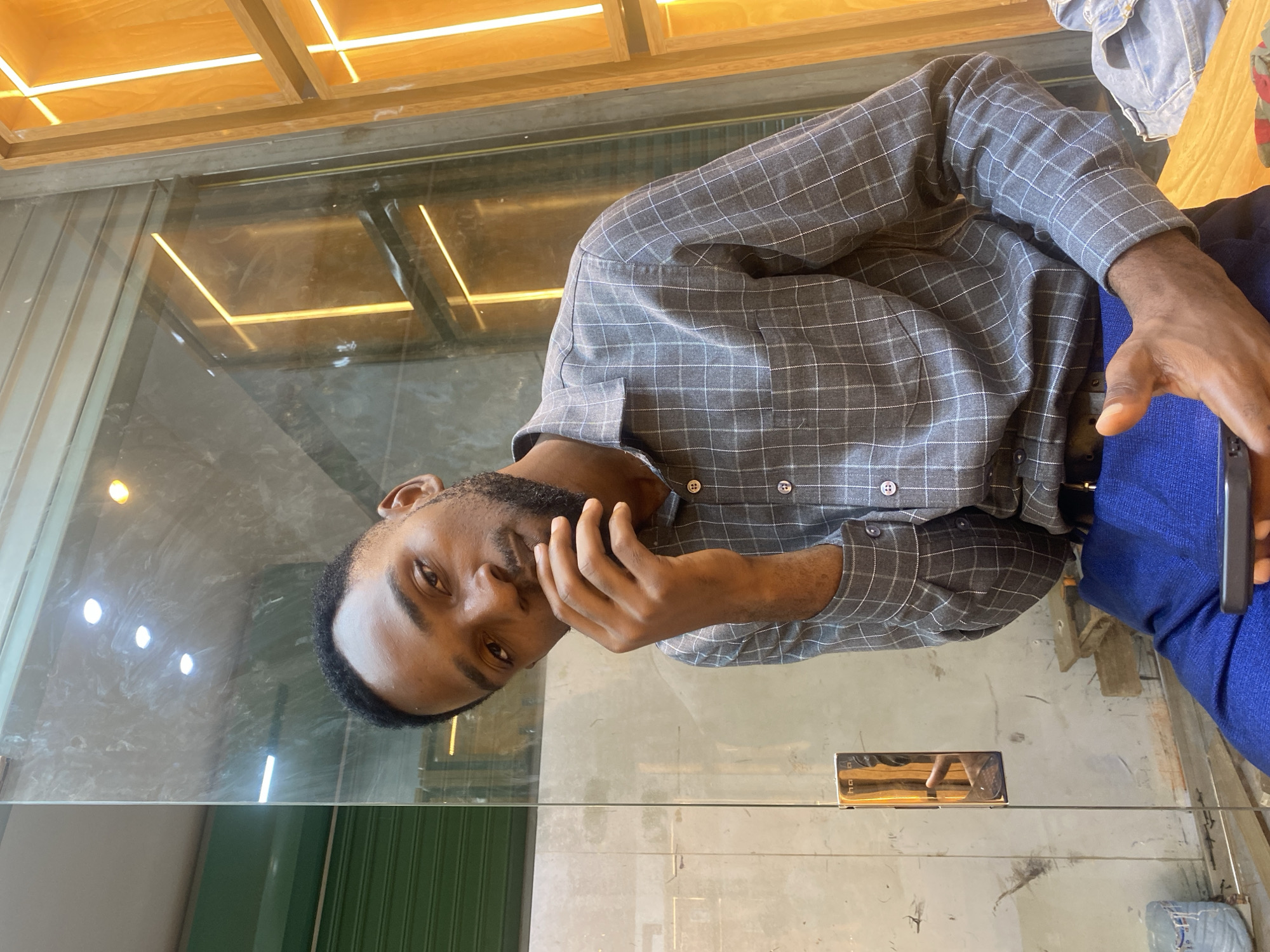 A man with dark skin and a beard is visible in the frame, wearing a grey plaid shirt and blue pants. He is holding a smartphone and has his hand near his face. The image appears to be taken through a glass surface, possibly a window or table, reflecting the environment. Overhead, there are wooden panels with linear light fixtures. The reflection on the glass surface reveals a cluttered background, possibly indoors, but the details are indistinct. There is no specific evidence provided to confirm that the location is in Dar es-Salaam, Tanzania.