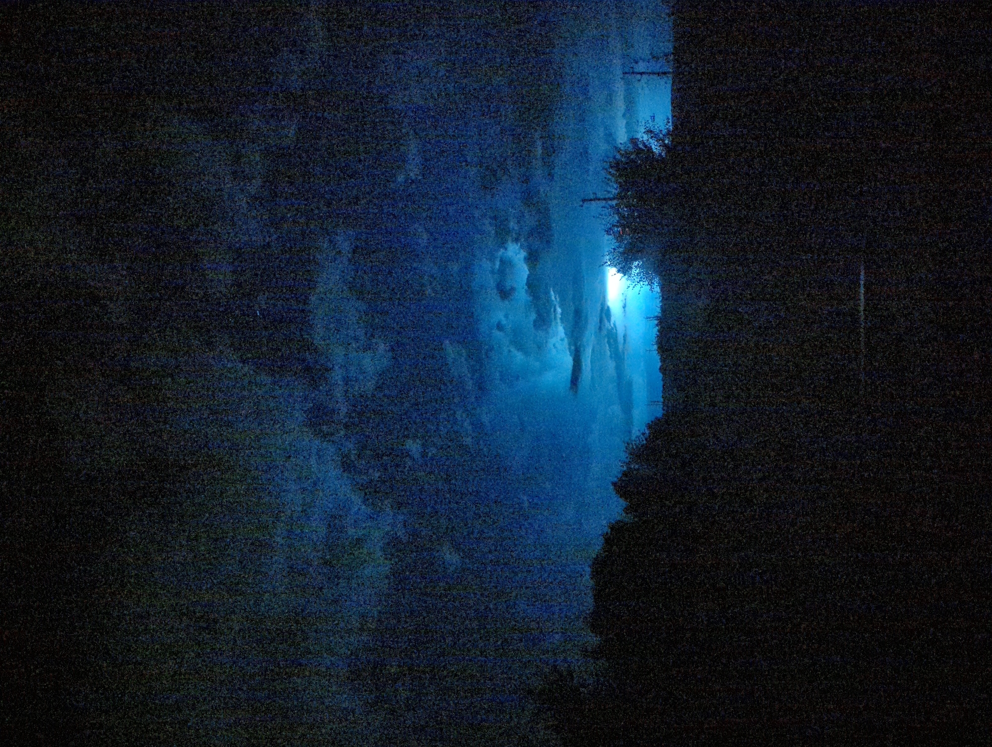 The image is a dark, grainy photograph taken at night or during twilight, depicting a natural landscape with a prominent sky. The sky is a deep blue, with textured clouds illuminated by a bright light source, likely the moon or a distant storm, visible in the center. Silhouetted against the sky are the dark outlines of trees and vegetation forming a low horizon line. The lower portion of the image is almost entirely black, obscuring any ground details. There are no visible people or specific man-made structures that could definitively place the location in Razgrad, Bulgaria, or any other city. The overall impression is one of a dark, atmospheric outdoor scene.