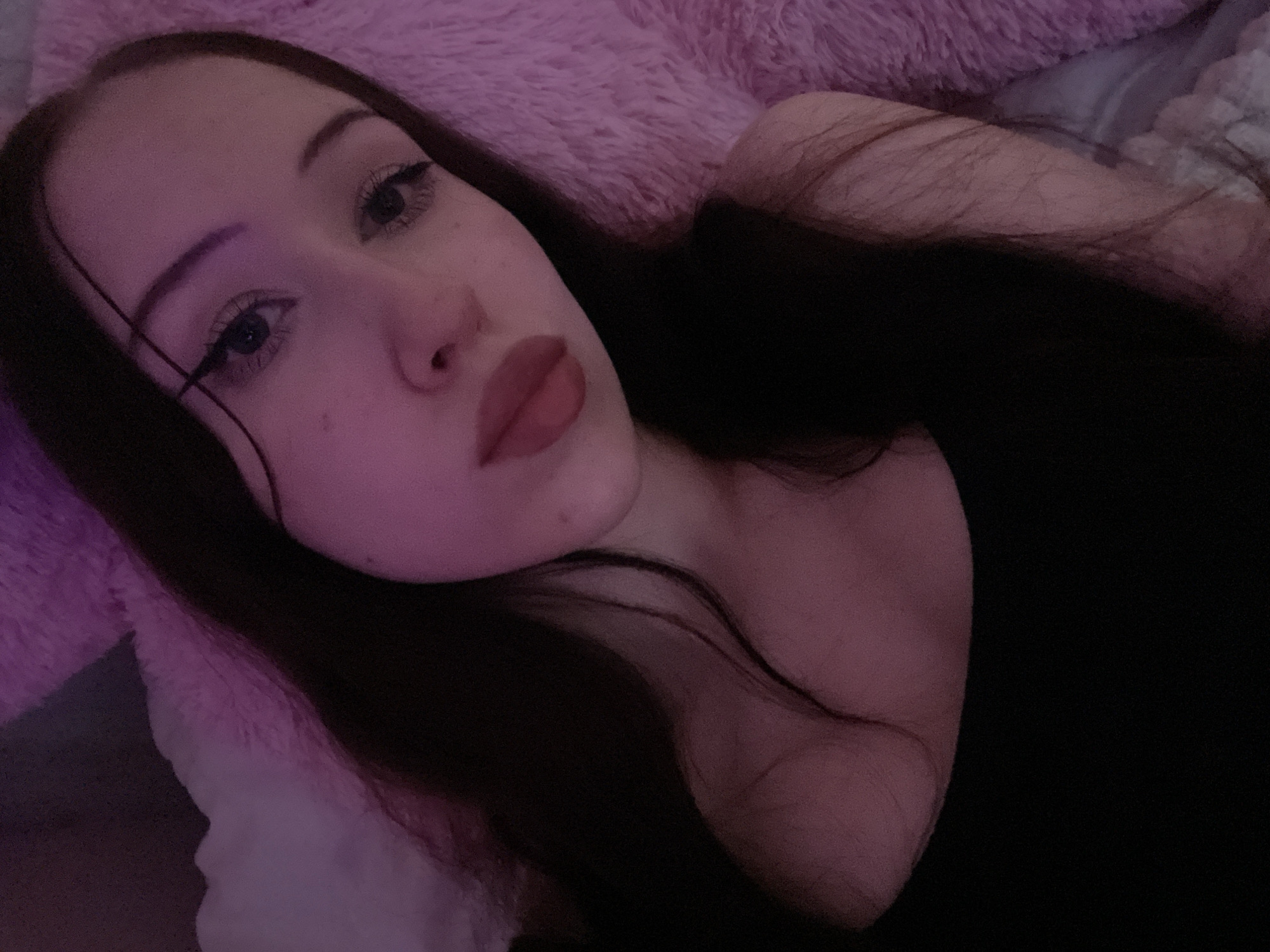 This media file contains a close-up, over-the-shoulder selfie of a young woman. She has long, dark hair and is wearing makeup, including eyeliner and lipstick. The woman is lying down on a fluffy pink surface, possibly a blanket or pillow, and the lighting is dim and has a pinkish hue, suggesting it's nighttime or the room is dimly lit. Her expression appears relaxed. There is no visible text. The context provided indicates this photo was taken in Votkinsk, Russia.