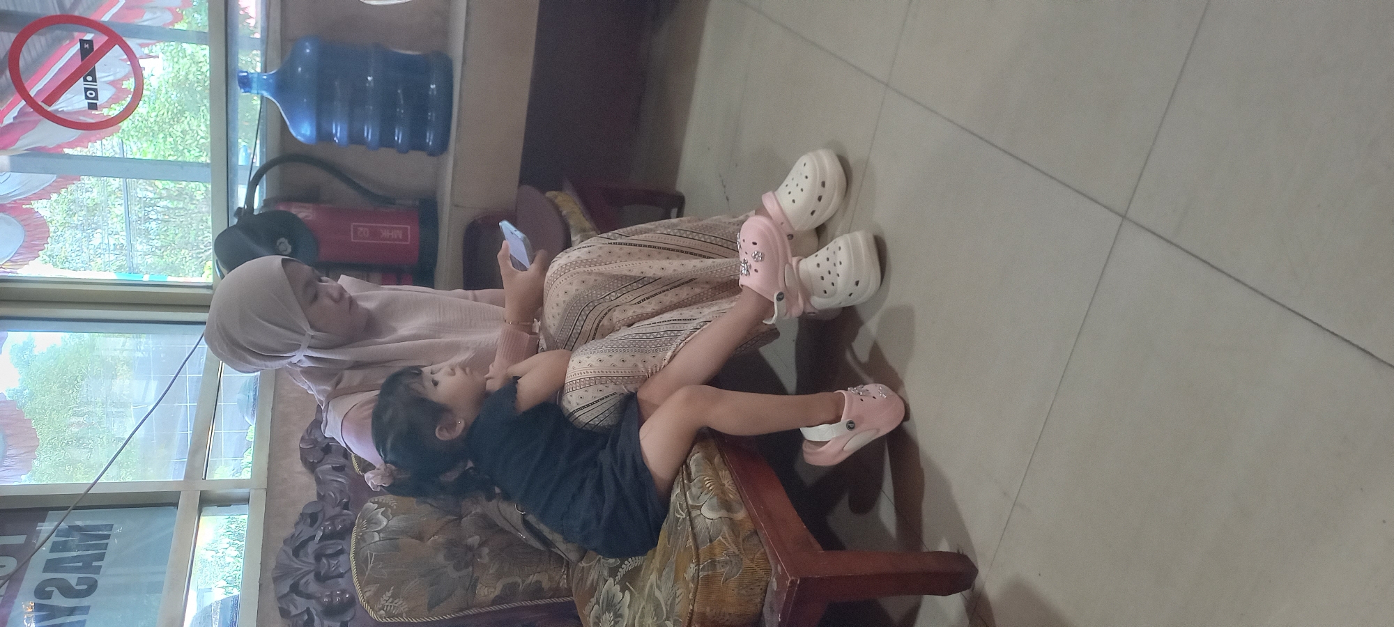 In Samarinda, Indonesia, two individuals, a young girl and an adult woman, are seated on a patterned armchair. The woman, wearing a light pink hijab and a matching patterned dress, is holding a smartphone. The young girl, dressed in a black garment, is seated on the woman's lap, her left leg extended forward and her right leg bent. She is looking to her right, with her left hand resting on her cheek. Both are wearing casual slip-on shoes; the girl is in pink, and the woman is in white. Behind them, a red fire extinguisher and a large blue water jug are visible. The background includes a window with greenery outside and a "no smoking" sign. The flooring is tiled in a neutral tone.