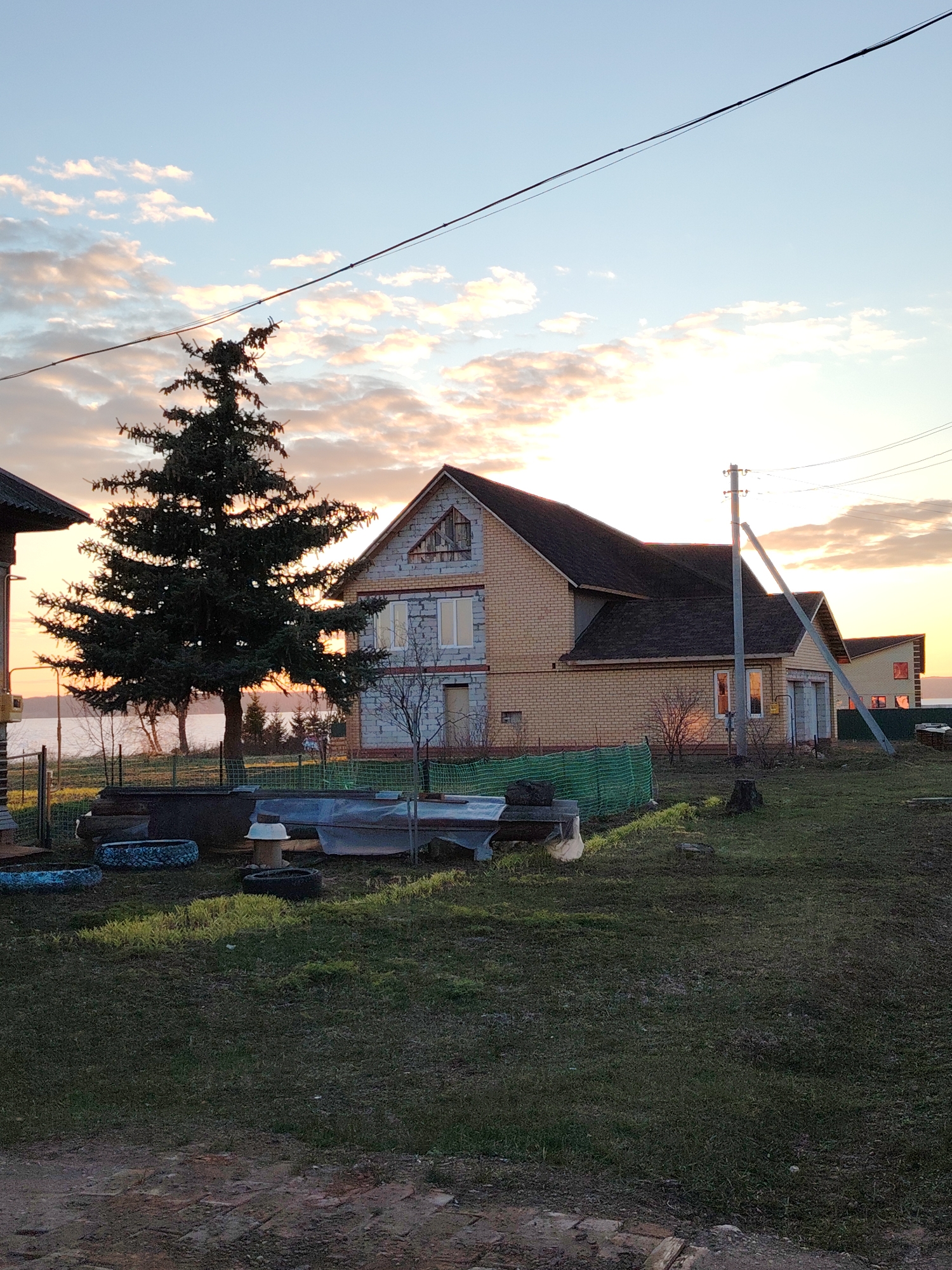 The image shows a two-story brick house under a sunset sky with scattered clouds, likely located in Elovo, Russia. An evergreen tree stands to the left of the house. A power line runs across the top of the frame. The yard has grass and some scattered objects, including what appear to be tires and possibly some garden equipment or supplies covered by a tarp. A power pole is visible near the house. The overall scene suggests a rural or suburban setting at dusk.