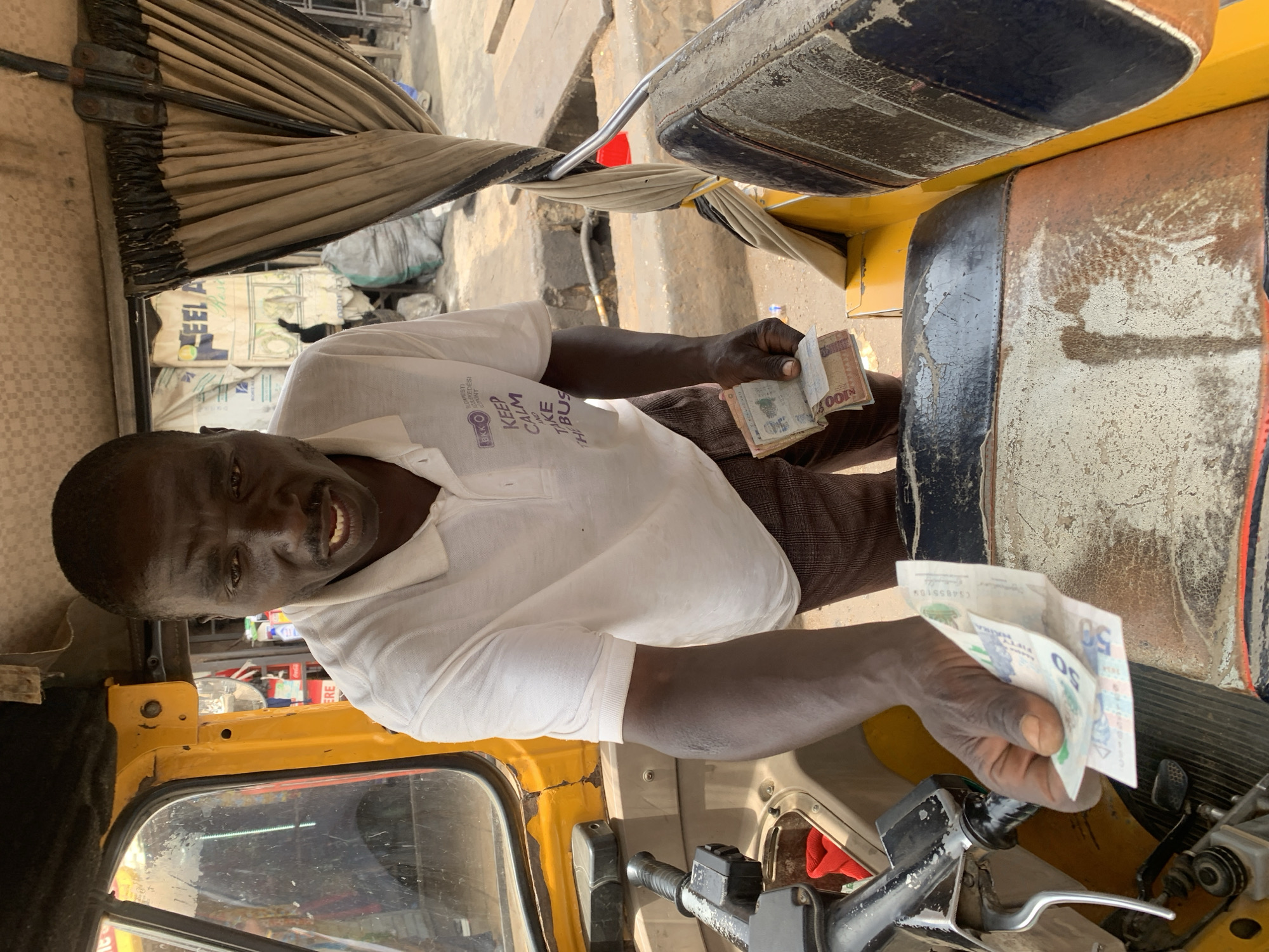 A man is sitting inside a yellow auto rickshaw in Jos, Nigeria. He is wearing a white collared shirt with a logo and words. He is holding Nigerian currency. One hand holds multiple 100 naira notes. The other hand holds multiple 50 naira notes. The vehicle's interior is worn. The seat is cracked and peeling. Outside the rickshaw is a dusty environment. There are large bags in the background. The location is Jos, Nigeria.