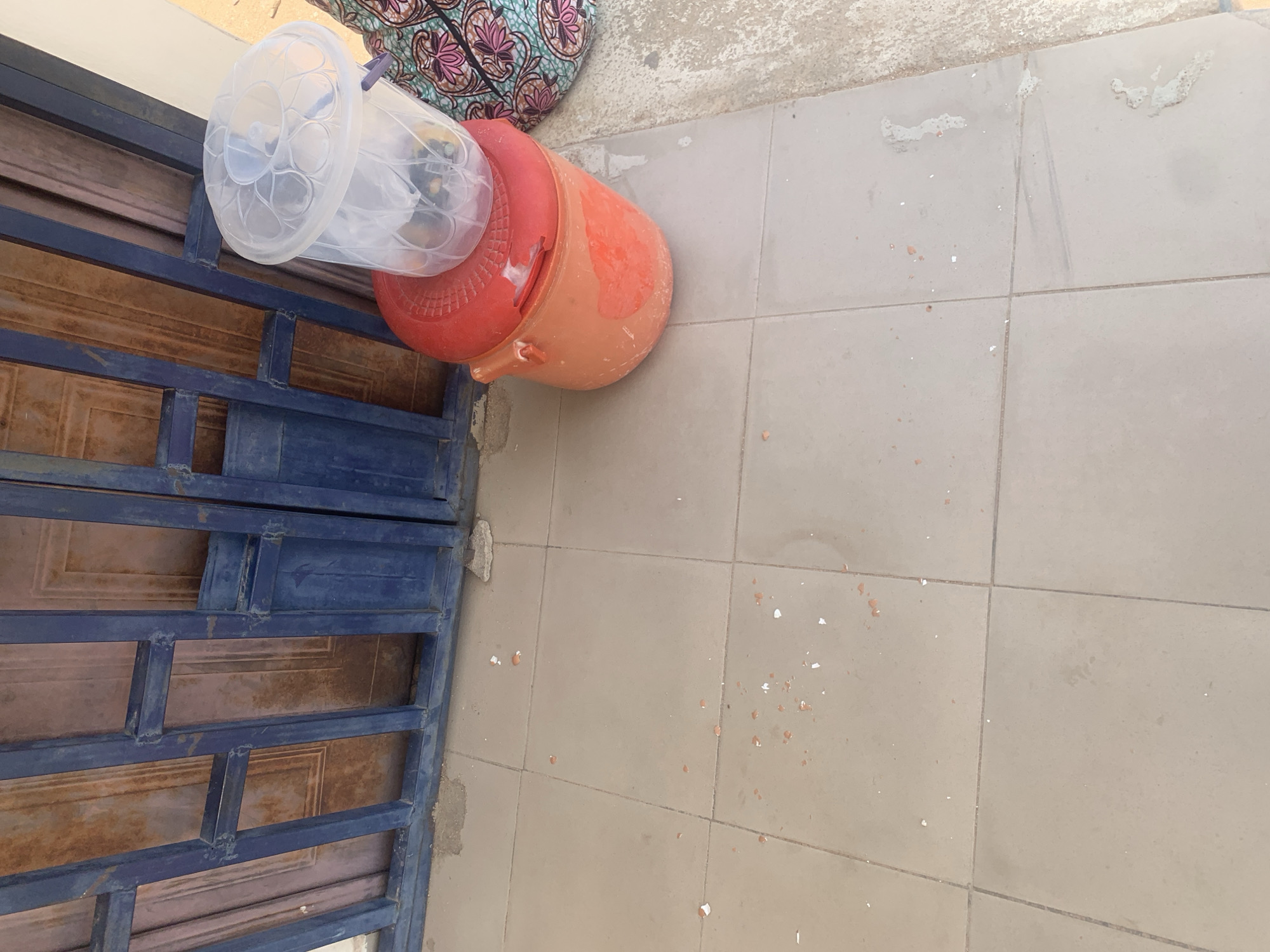 In Jalingo, Nigeria, an outdoor tiled surface is observed. A blue metal gate with ornate detailing stands in the background. In the foreground, an orange plastic bucket is present, topped with a clear plastic lid. Inside the clear lid, several yellow objects, possibly fruits, are visible. Adjacent to the bucket, a piece of fabric with a pink and teal floral pattern is partially visible. The tiled floor shows scattered debris, including small white and reddish-brown fragments, likely from broken items.