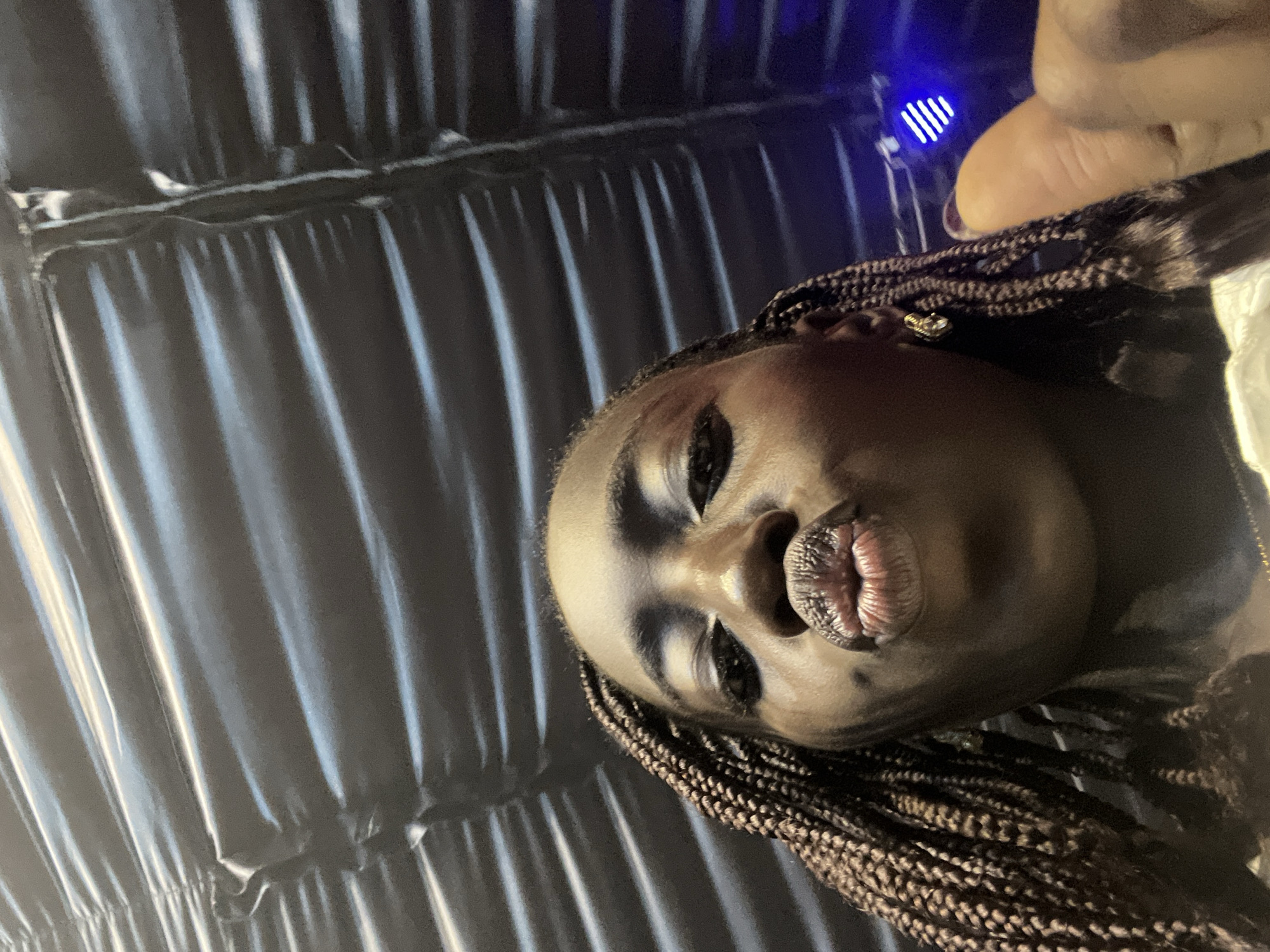 A close-up, low-angle selfie features an adult female with dark skin and dark braided hair, facing the camera with her lips pursed. She wears small, gold-toned stud earrings. Her right thumb, exhibiting dark nail polish, is visible in the lower right corner, likely holding the camera. The background comprises a dark, reflective, and horizontally corrugated or pleated surface, possibly a ceiling or inflatable structure. A distinct blue LED light source is visible in the upper right background. The scene is situated in Galadimawa Village, Nigeria, under dim, artificial lighting.