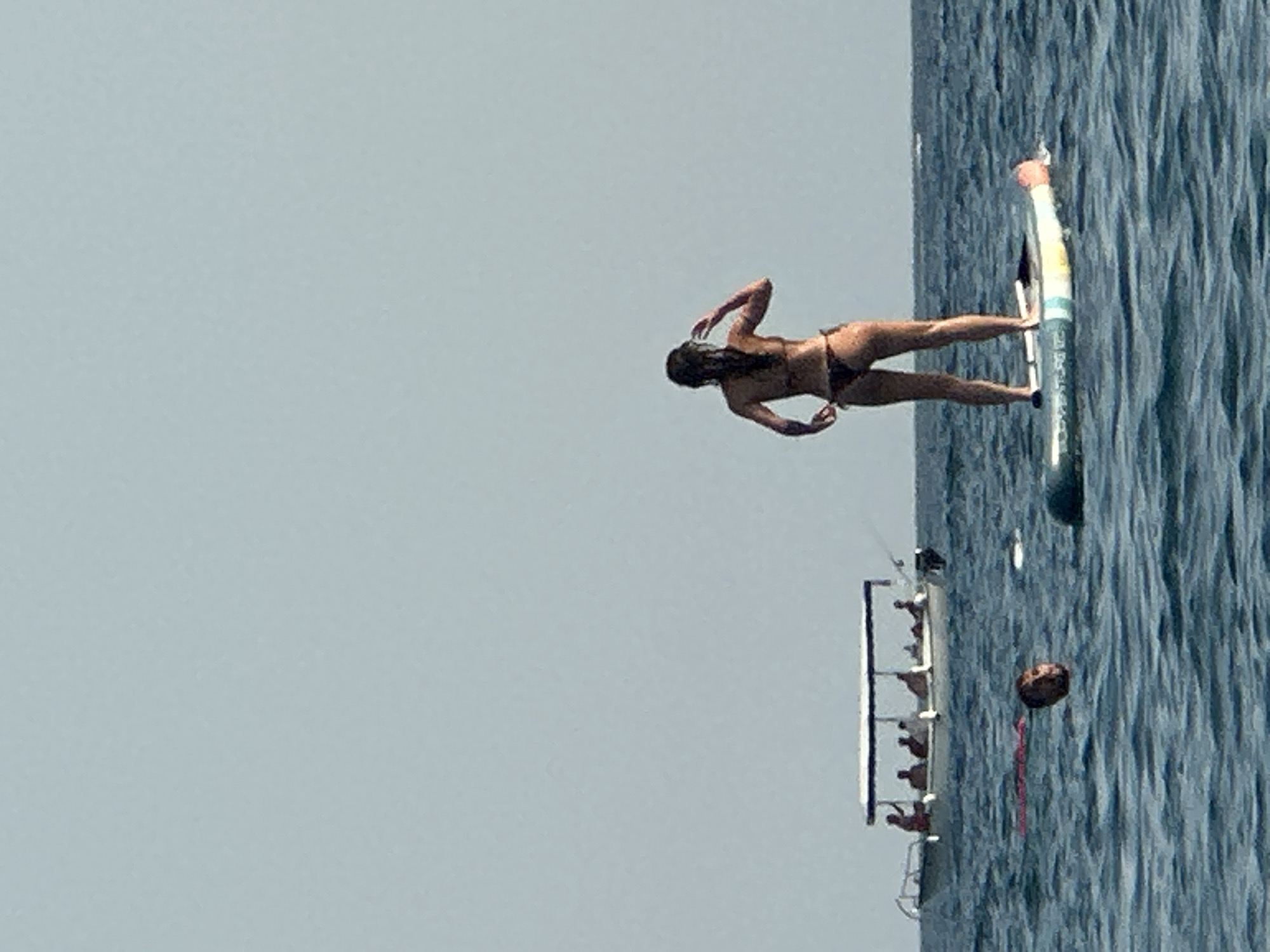 In Boreti, Montenegro, a woman in a bikini stands on a paddleboard in the ocean. She is facing away from the camera, with her right hand touching her hair. Her paddleboard is positioned in the foreground, and the water is choppy. In the background, a boat carrying several people is visible on the water. To the left of the boat, a person's head is visible above the water. The sky is clear and blue.