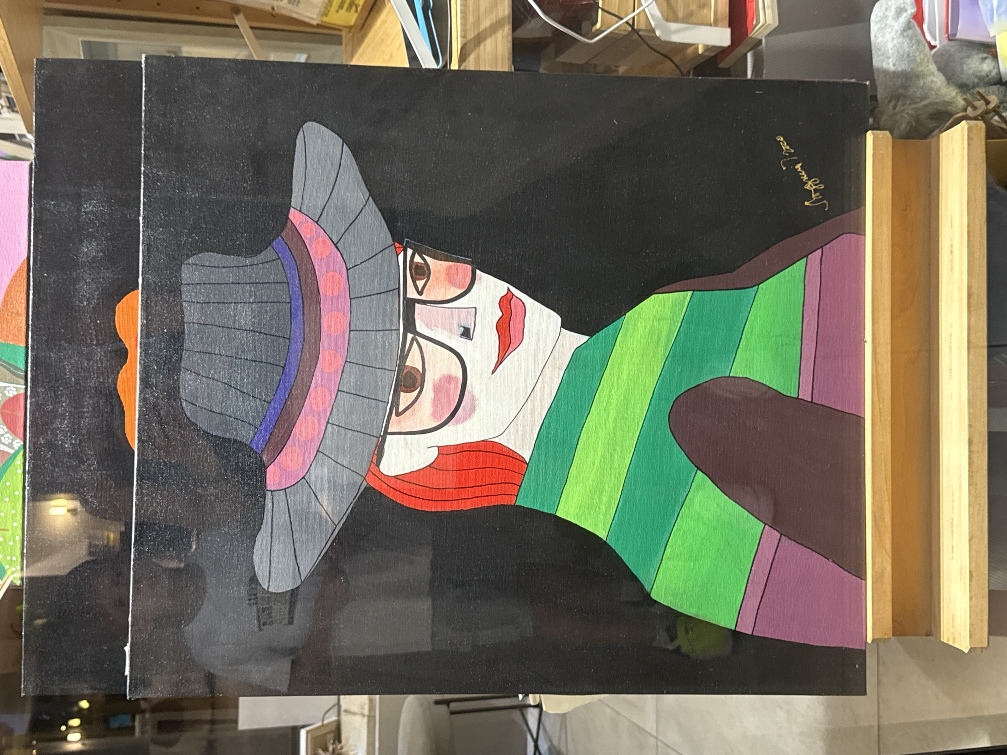 The image shows a painting of a stylized woman wearing a wide-brimmed hat and glasses. The woman has red hair and is depicted with a green and purple striped garment. The background of the painting is black, and there is a signature in gold script in the lower right corner. The painting is displayed on a wooden easel. In the background, through a glass reflection, a person in a white t-shirt can be seen. This scene is likely located in Budva, Montenegro, given the context provided.