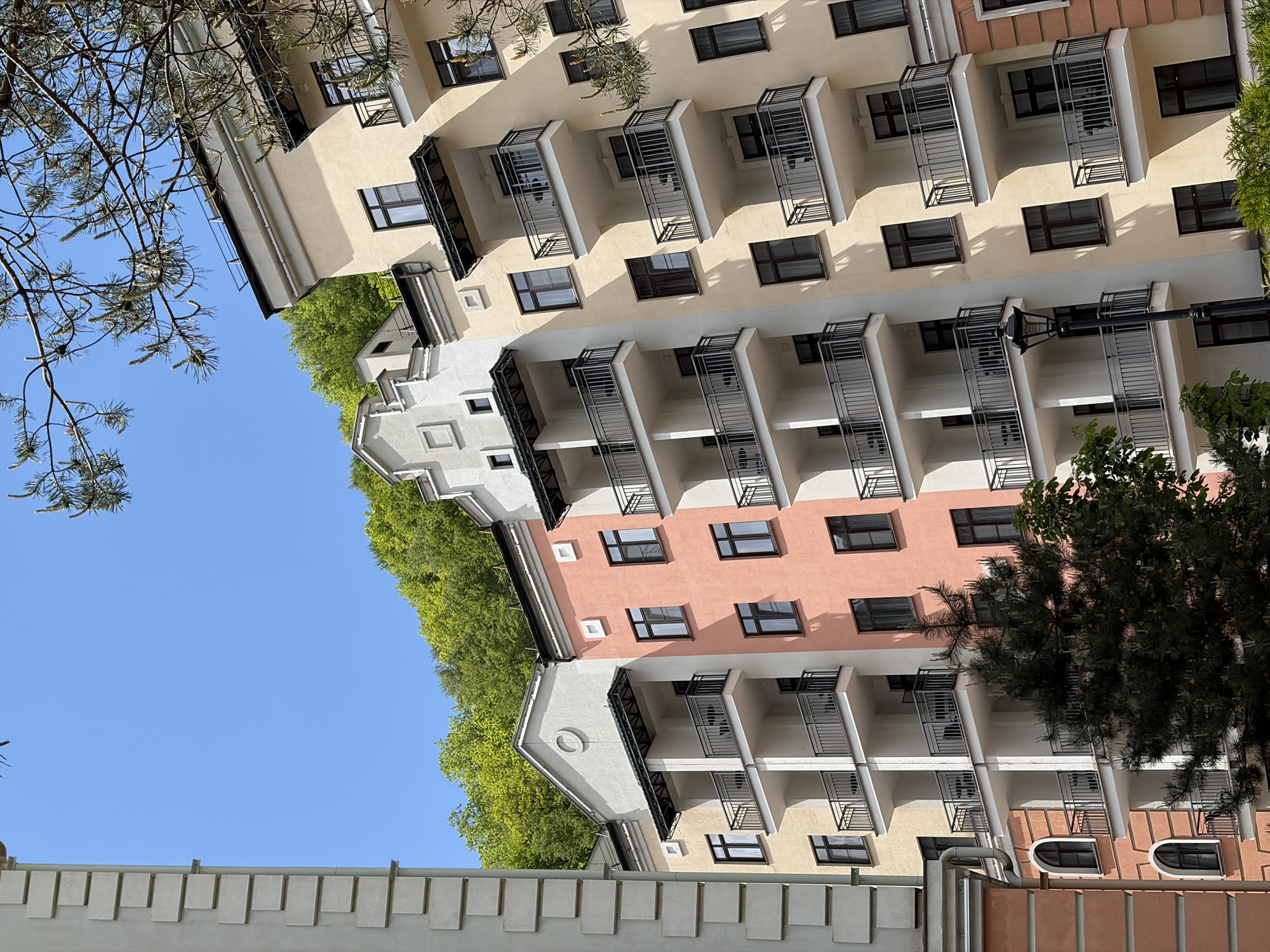 The image captures a multi-story building in Estosadok, Russia, observed from a low angle. The building features a color scheme of cream, white, and pink, with prominent balconies and dark-framed windows. Vegetation, including trees and greenery, flanks the building's edge. The sky is visible in the upper left, appearing clear and blue. A section of a low concrete wall is visible in the lower part of the frame.