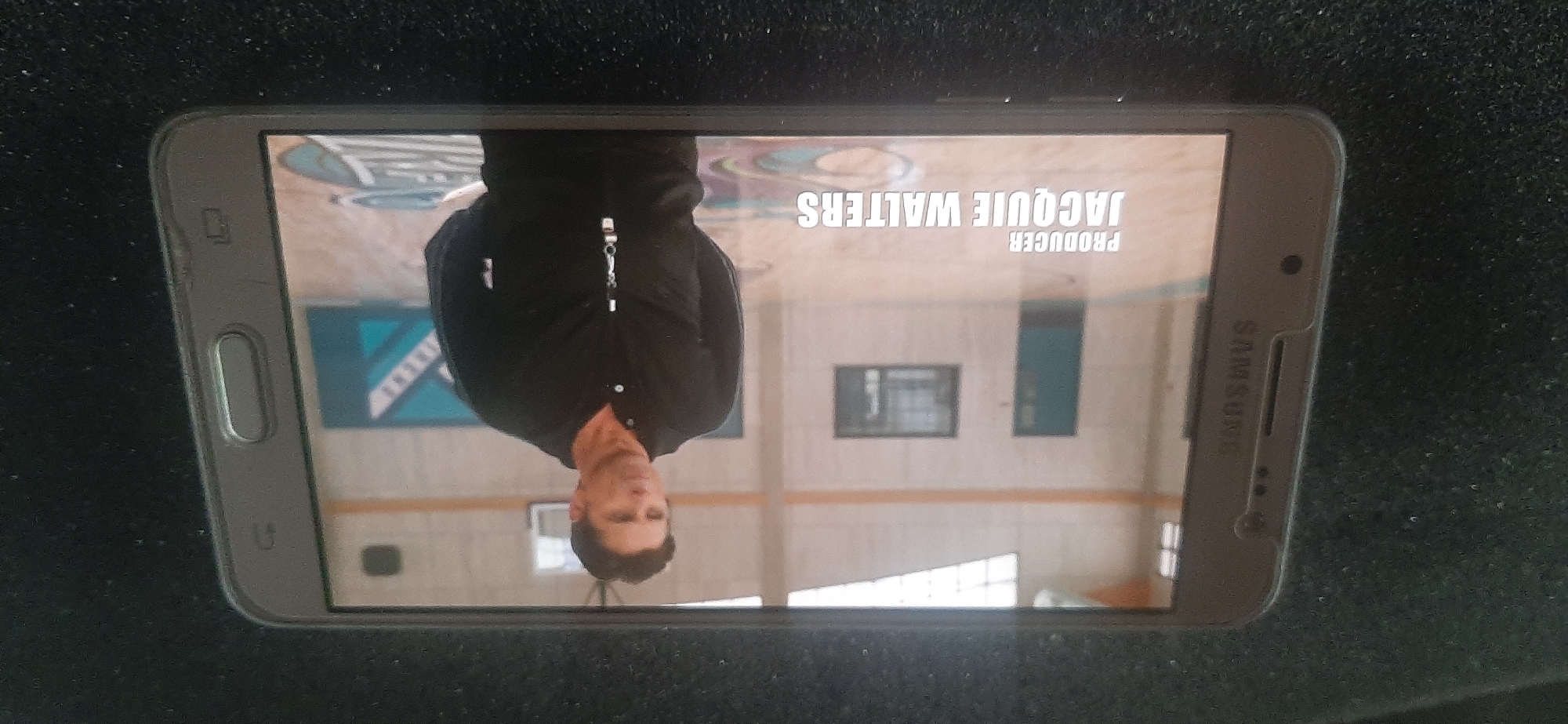 The image displays a smartphone screen showing a scene from a television show or movie. On the screen, a man wearing a black collared jacket is standing in a gymnasium. Text overlay on the screen reads "PRODUCER JACQUIE WALTERS". The smartphone itself is dark gray and is placed on a dark, textured surface. The image does not contain any information about the city of Yessentukskaya, Russia.