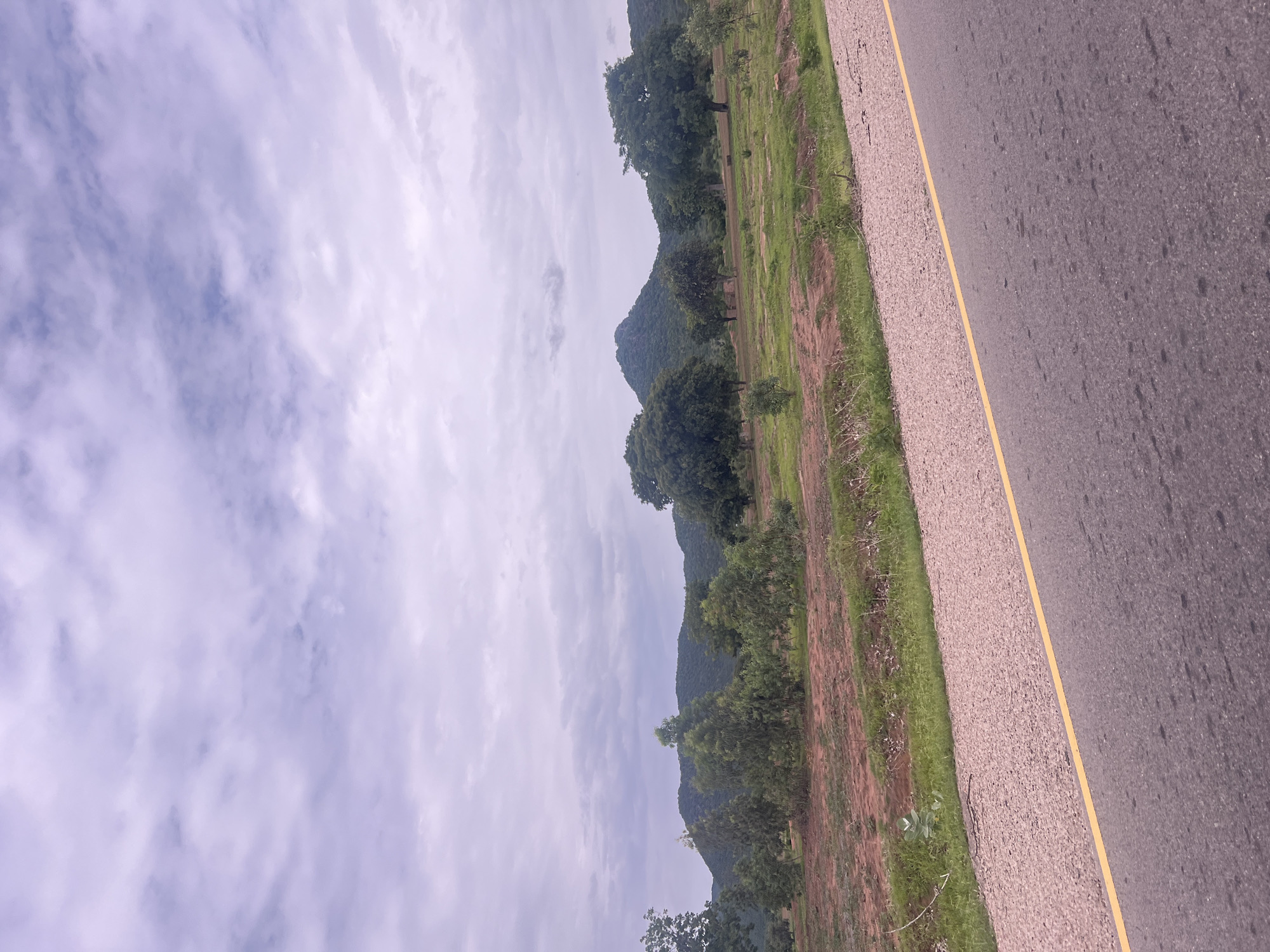 The image depicts a roadside view in Minda, Nigeria, on a cloudy day. The scene is dominated by a paved road on the right side, marked by a yellow line along its edge. To the left of the road is a strip of gravelly shoulder, followed by dry, reddish-brown earth and patches of green grass. Further to the left, a line of trees and vegetation stretches towards a backdrop of rolling hills covered in dense green foliage. The sky is overcast with soft, white clouds that obscure the sun, suggesting it could be morning or late afternoon. There are no people or discernible objects other than the road and natural elements. The overall mood is serene and natural, conveying a sense of rural landscape.