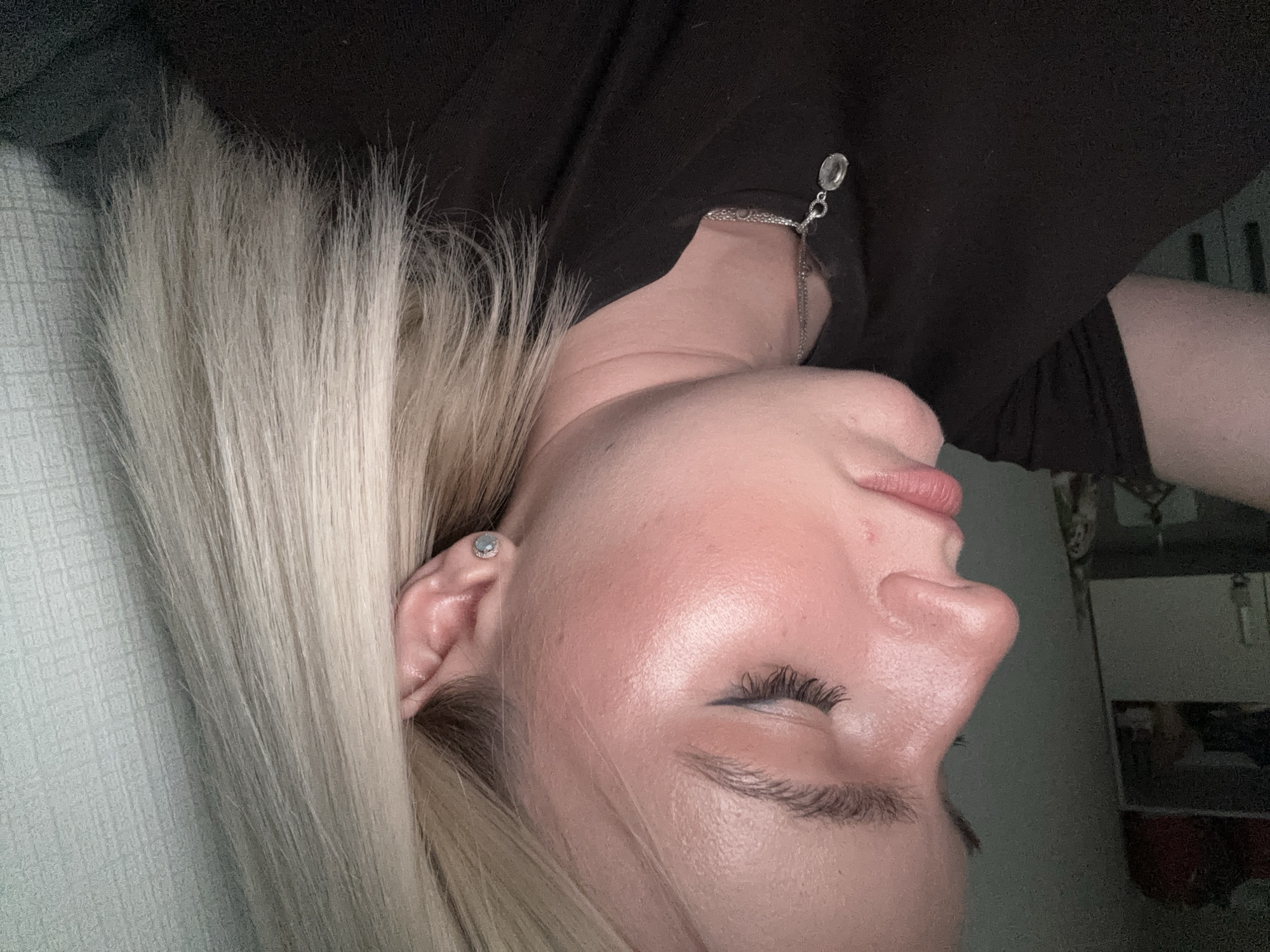 The image shows a woman with platinum blonde hair, wearing a black top and a silver chain necklace. She has fair skin, a closed eye with eyeliner, and an earring. The background is blurry, showing a light-colored wall. There is no information to suggest that the image was taken in Vsevolozhsk, Russia.