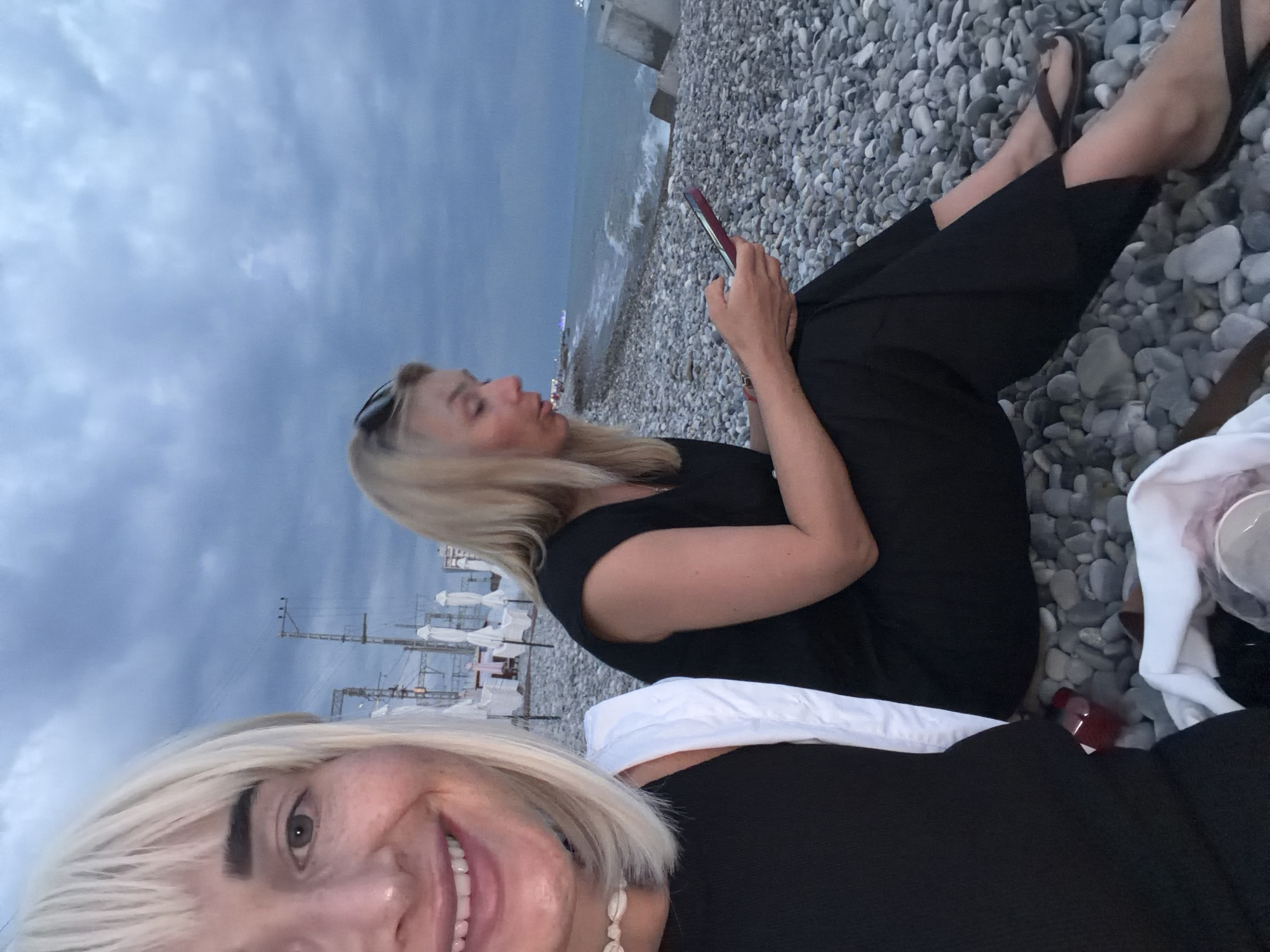 Two women are sitting on a pebble beach near the ocean at dusk. The woman in the foreground, on the left, is taking a selfie and smiling. She has blonde hair and is wearing a black top with a white strap and a seashell necklace. The woman in the background, on the right, is looking at her phone and wearing a black dress. The sky is cloudy, and the ocean is visible in the background with waves breaking on the shore. There are also some white umbrellas and structures visible on the beach in the distance.