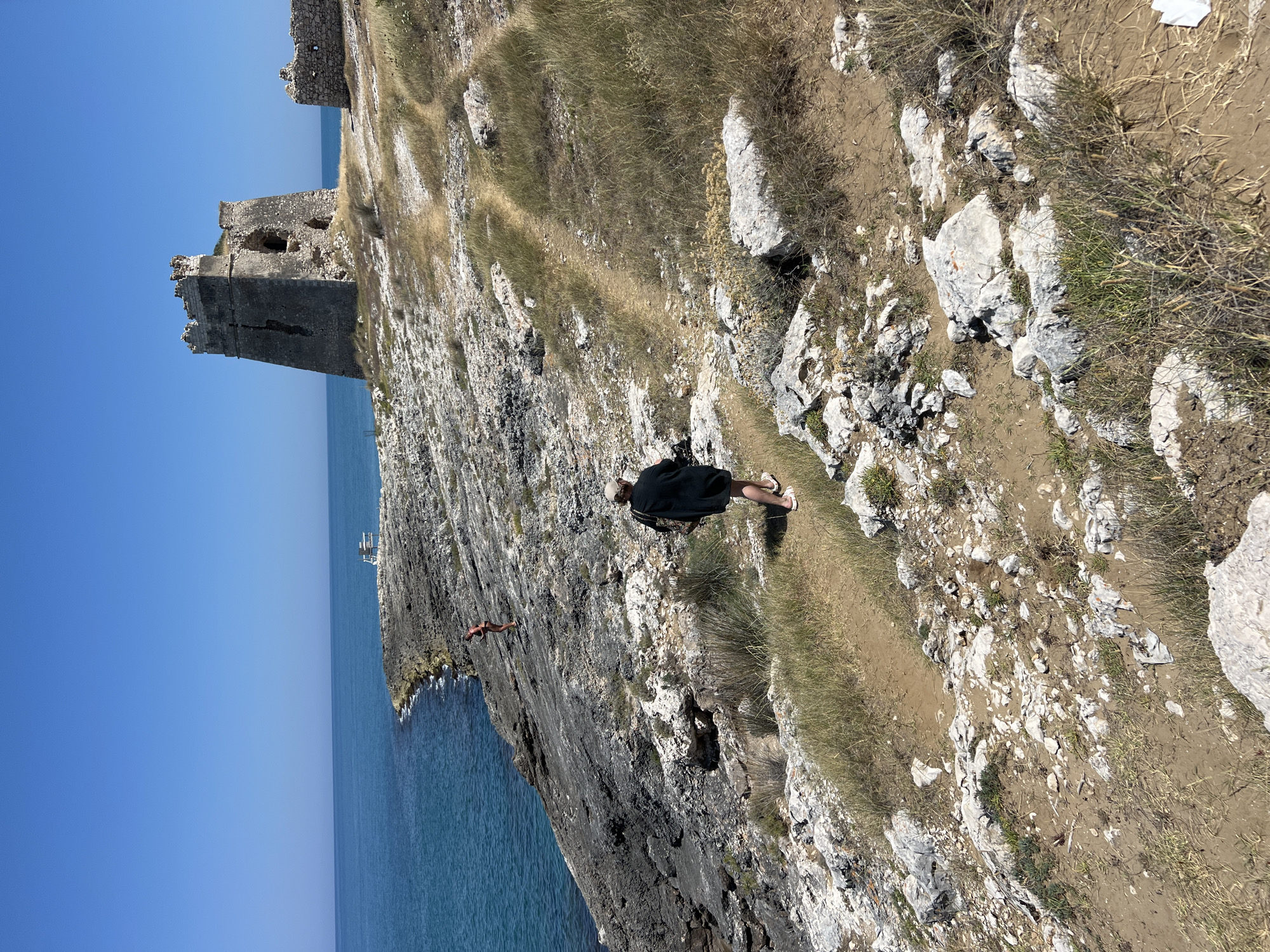 A person in a black shirt and beige shorts stands on a dirt path near a cliff overlooking the sea. The path is lined with dry grass and rocks. Further down the cliff, another person in red shorts is visible near the water's edge. In the background, the ruins of a stone structure, possibly a castle, perch on the cliff. The sky is a clear, bright blue, suggesting a sunny day. The sea is a deep blue and calm. The scene appears to be a scenic coastal location, likely a historical site given the ruins. The overall impression is one of exploration and appreciation of natural beauty and history.