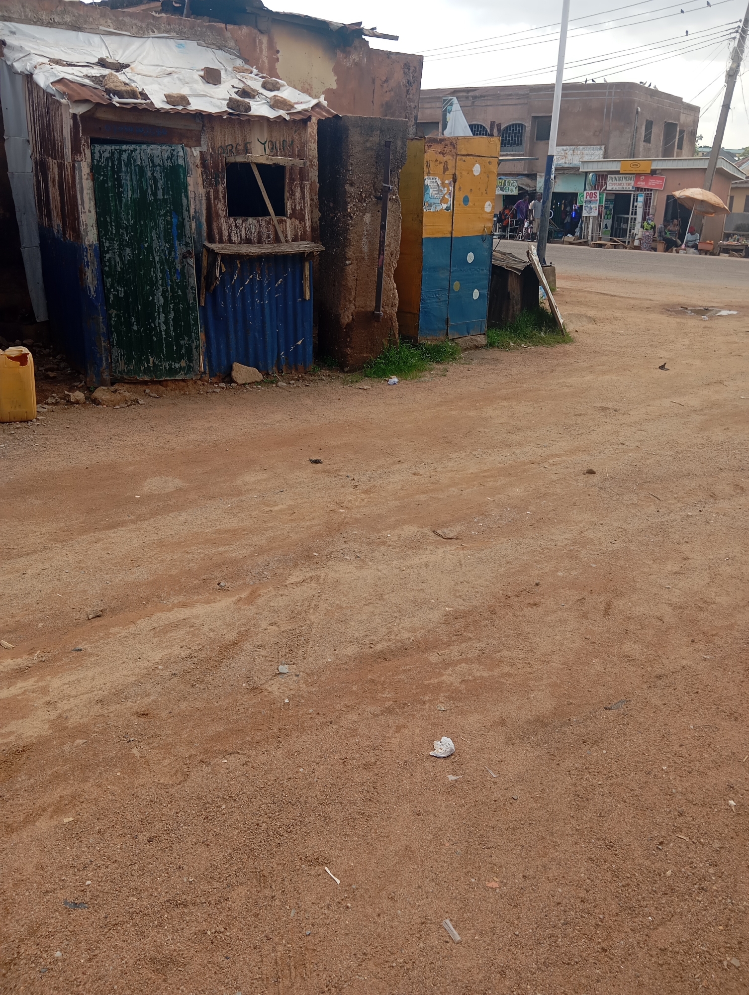The image depicts an outdoor scene in Jos, Nigeria. In the foreground, a sandy, dirt road takes up a significant portion of the frame. Scattered on the road are small pebbles, debris, and a few pieces of white paper.
To the left of the frame is a dilapidated, makeshift structure. It appears to be constructed from corrugated metal sheets, some of which are rusted. The structure has a green-painted door and a window opening with wooden bracing. Blue corrugated metal is visible on the lower half of the structure. A yellow plastic container sits next to the structure. Above the window, text is visible, though partially obscured and difficult to read. Faintly legible words appear to be "FREE YOUR..."
Adjacent to this structure, and slightly to the right, are two tall, colorful boxes, stacked one on top of the other. The top box is yellow with some white circular patterns, and the bottom box is blue with similar white circular patterns. These boxes are positioned in front of a concrete pillar and a building.
In the background, a street scene unfolds. Several buildings are visible, some appearing to be shops. One building has a sign that reads "ANNEBOGH COMMERCIAL AGENCY." Another sign is partially visible, possibly indicating a "POS" service. People are seen in the background, some gathered near the shops, and one person is seated under a beige umbrella. A white pole, likely a utility pole, stands tall in the middle ground, with electrical wires extending from it. The sky is overcast, suggesting a cloudy day. The overall impression is of a modest, possibly commercial, area in a town or city.