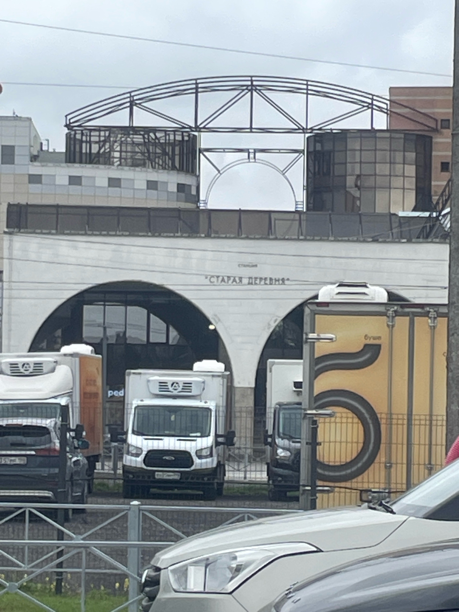 The image shows a cloudy day in front of a building with arched entrances. Above the arches, text reads "СТАНЦИЯ" and "СТАРАЯ ДЕРЕВНЯ". There are several vans and trucks parked in front of the building. One van is white with a refrigerated unit on top. Another vehicle is a dark SUV. A yellow truck with a large black "5" graphic is also visible. In the foreground, a silver car is partially visible. The scene appears to be outdoors, possibly in a commercial or transportation hub.