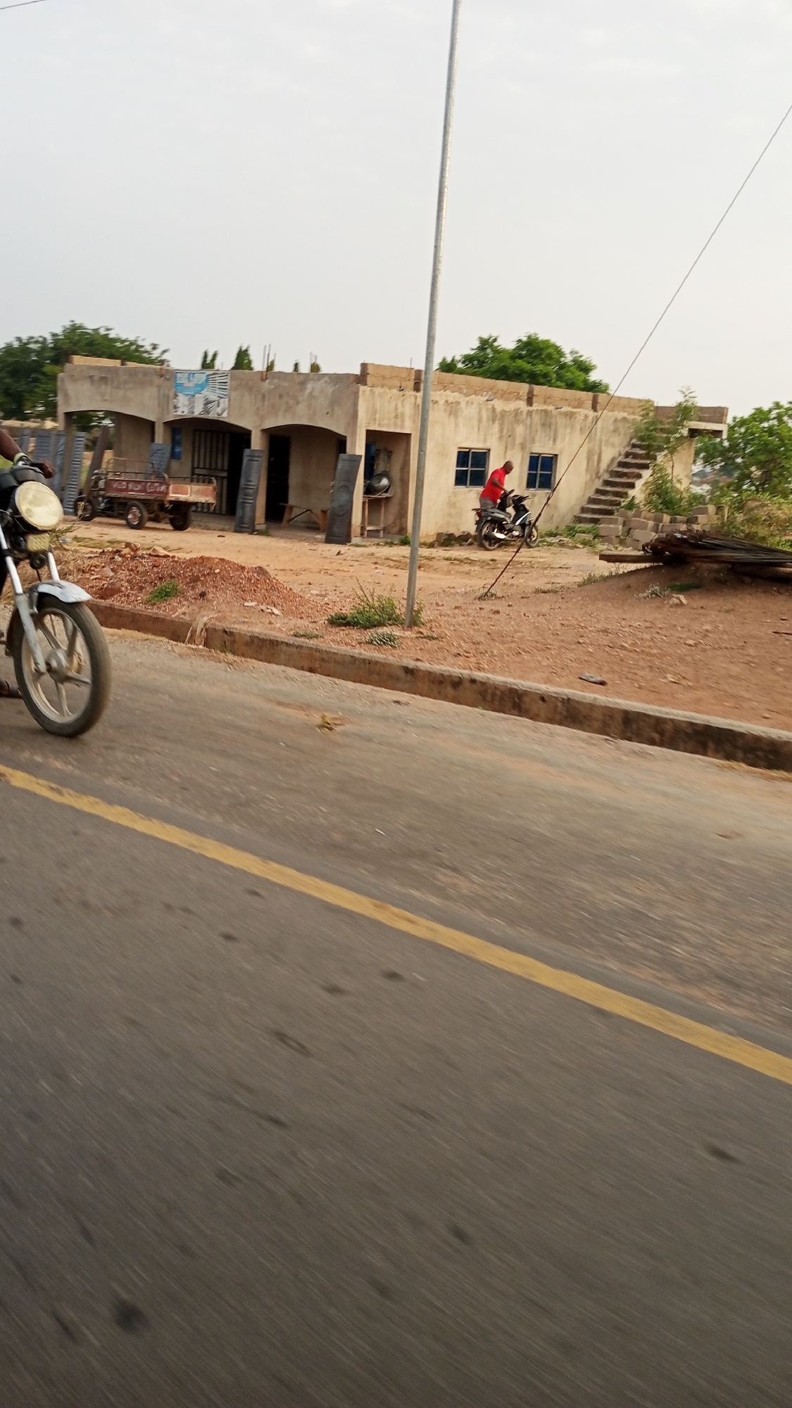 The image shows a roadside scene in Zango-ndaji, Nigeria. A building with unfinished concrete construction and arched entrances is visible in the background. Two motorcycles are present, one partially obscured in the foreground and another with a person in a red shirt riding it near the building. A yellow line marks the edge of the asphalt road in the foreground. A utility pole stands tall near the building. The surrounding area is characterized by bare earth and scattered objects, including construction materials.