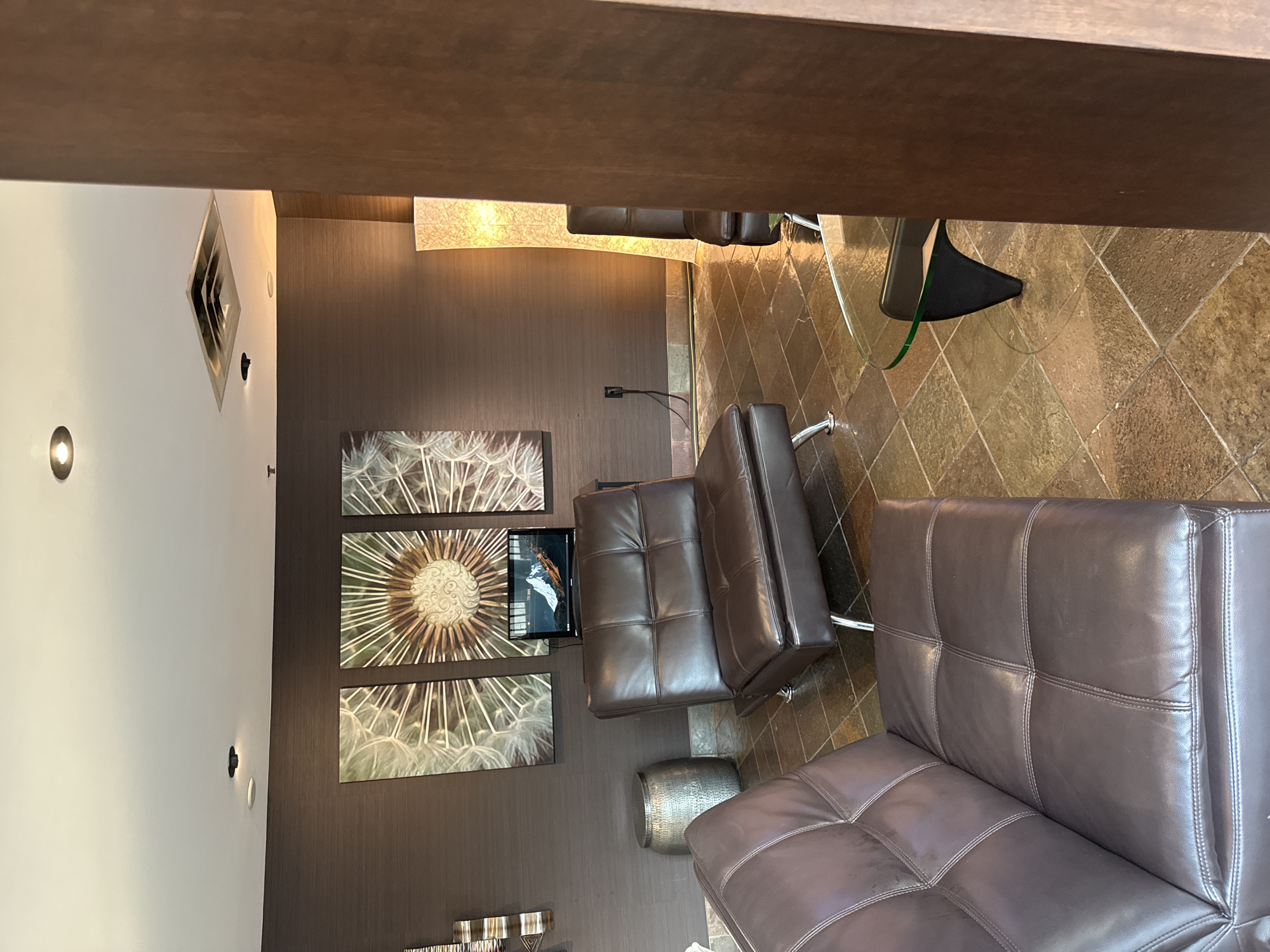 This is an indoor image of a waiting area or lounge, likely located in North Vancouver, Canada. The room features a dark brown textured wallpaper and a tiled floor in earthy tones. In the foreground, a dark brown leather sofa is visible. Further back, another dark brown leather sofa and a matching armchair are positioned around a glass-topped coffee table with a dark base. On the wall, three large framed artworks depict dandelions. A flat-screen monitor sits on a stand between two of the dandelion artworks, displaying an image. A tall, illuminated floor lamp stands to the right of the armchair, casting a warm glow. A metallic cylindrical stool is placed to the left of the sofa. The ceiling has recessed lighting and an air vent. The scene is static and appears to be unoccupied.