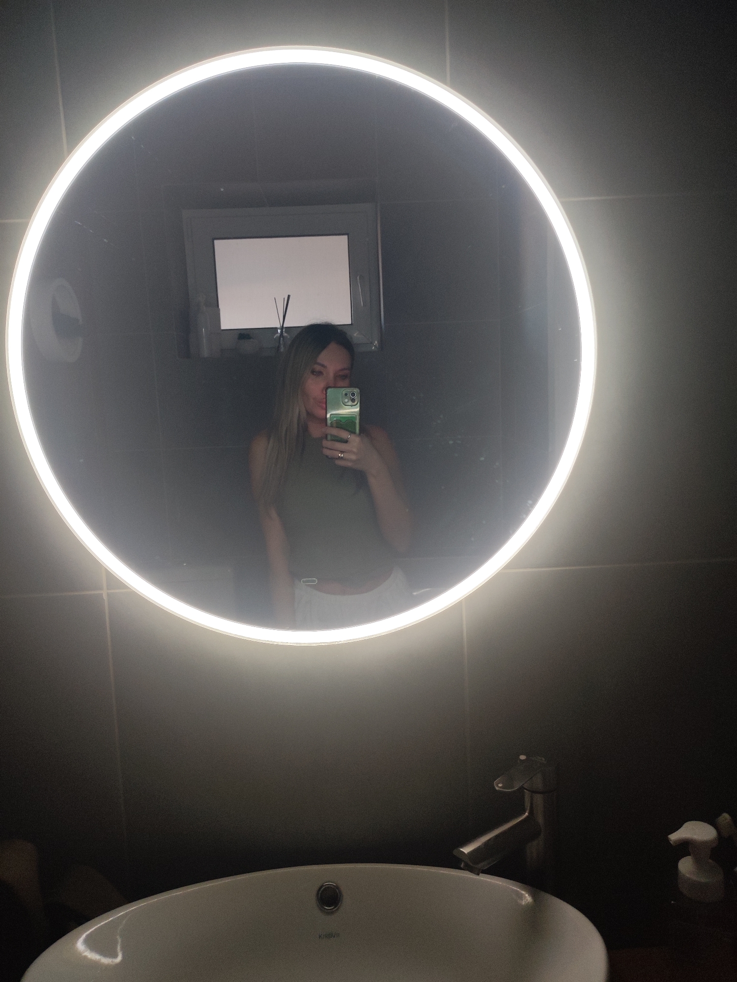 A woman is taking a selfie in a bathroom mirror. The mirror is round with a built-in light. She is wearing a green top and white bottoms. The bathroom has gray tiled walls, a white sink, and a silver faucet. There is a window above the mirror and a soap dispenser on the counter. The location is not discernible and therefore the city of Votkinsk, Russia cannot be confirmed.