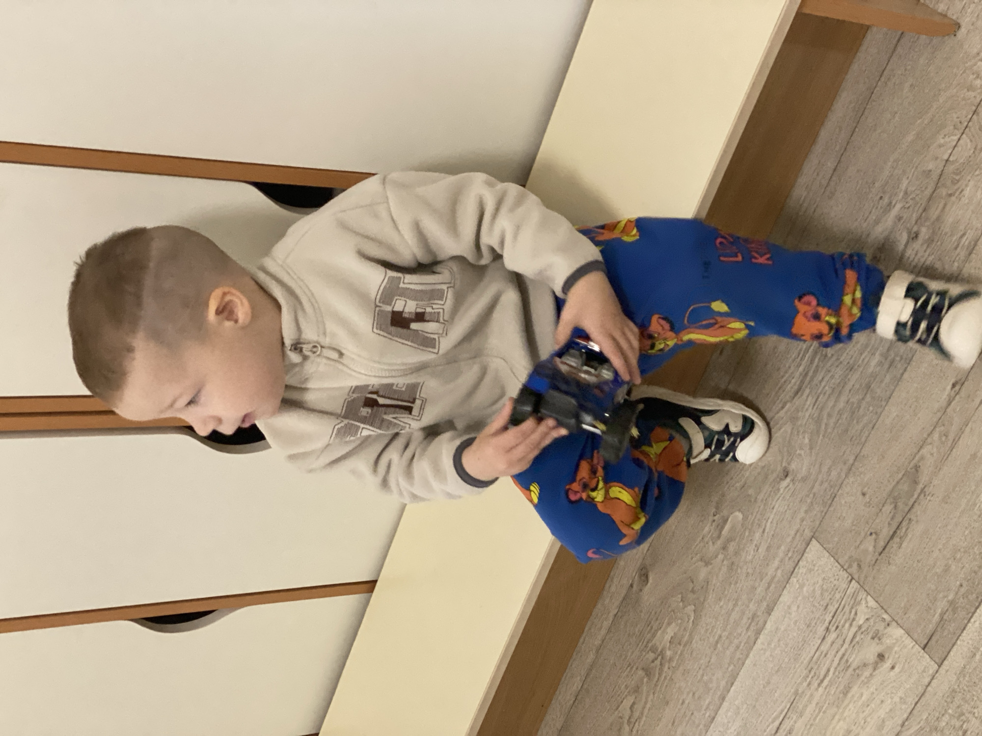 A young boy is sitting indoors playing with a toy car. He is wearing a light beige zip-up sweatshirt with an "ET" logo, blue Lion King patterned pants, and black and white sneakers. The boy is sitting on a beige surface next to a set of white drawers with brown trim. The floor is light gray with a wood grain pattern. The location is in a building in the city of Sochi, Russia.