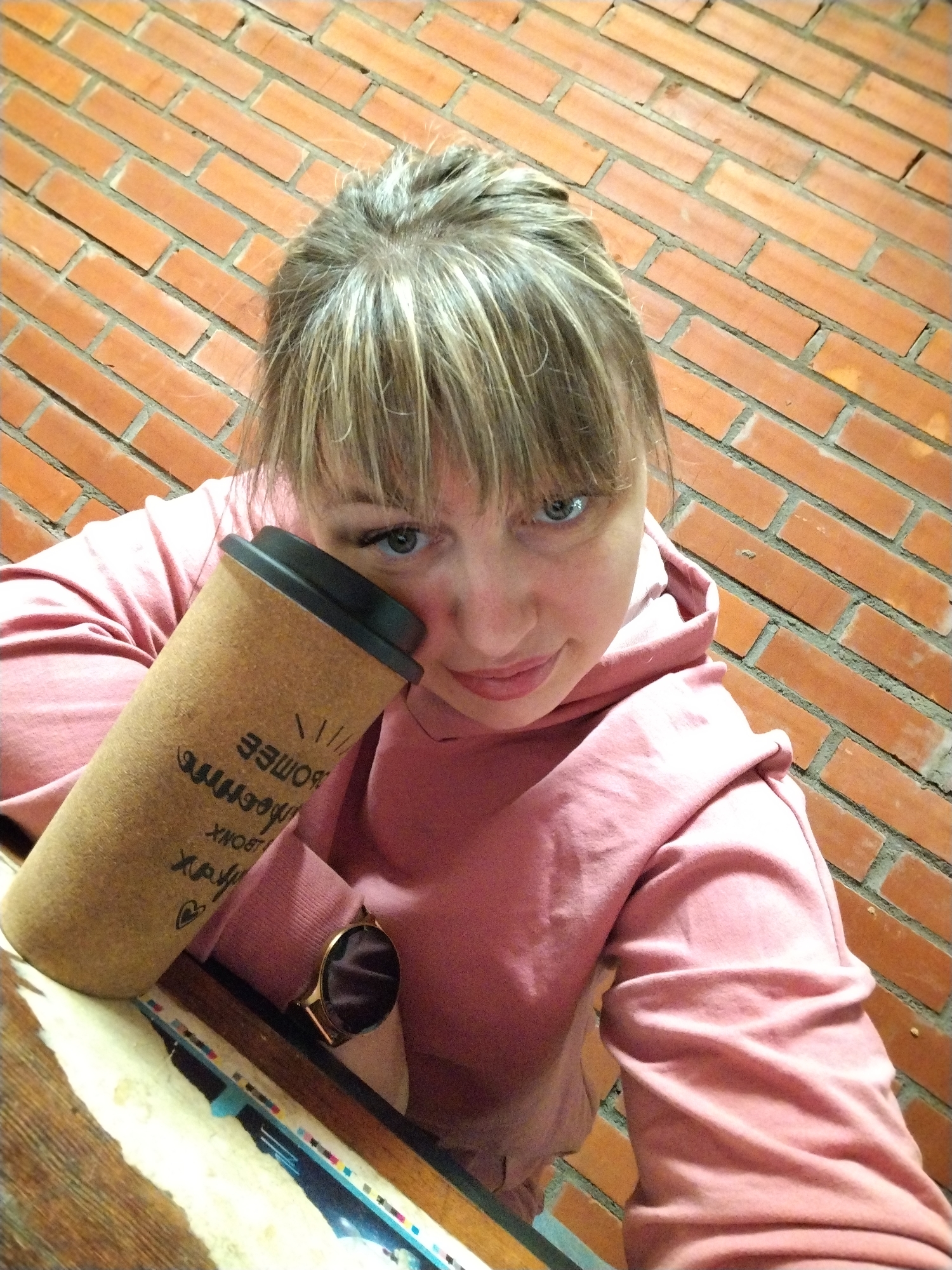 A young woman, likely in her 20s or 30s, is captured in a close-up selfie, looking directly at the camera with a subtle, gentle smile. Her light-colored hair, possibly blonde or light brown with highlights, is styled with bangs, framing her bright blue or green eyes. She wears a pink or dusty rose-colored hooded sweatshirt.
She holds a reusable coffee cup, appearing to be made of cork or a similar textured material, with a black lid. She rests her head slightly against the cup, which features handwritten text in Cyrillic, reversed due to the selfie perspective. Partially legible words include "БОЛЕЕ" (meaning "more") and "ПРОЧНОЕ" (meaning "durable" or "strong"), alongside "БЛОНК", a word starting with the Cyrillic letter 'Х', a small heart symbol, and "///". On her left wrist, a smartwatch with a gold-colored bezel and a dark screen is visible.
The background consists of a rustic red brick wall, suggesting an indoor or sheltered outdoor setting such as a cafe or a cozy room. A wooden surface, possibly a table, is visible in the foreground. The woman's expression conveys a calm and content mood. The image provides no specific time of day or weather cues. The location context provided is Заволжье, Russia.
