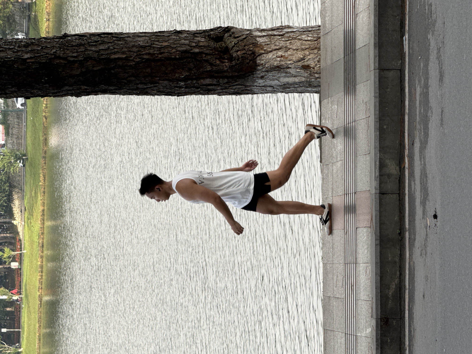 A young Vietnamese man, dressed in a white tank top and black shorts, jogs along a paved walkway beside Xuan Huong Lake in Da Lat, Vietnam. He is moving from left to right across the frame, his arms swinging naturally with his stride. The walkway is bordered by a low concrete curb and the rippling surface of the lake. To the right of the man, a large, textured tree trunk rises vertically, its bark dark and weathered. In the background, across the lake, a lush green park area is visible with trees and some distant figures. The overall scene depicts a peaceful moment of exercise in a scenic urban setting.