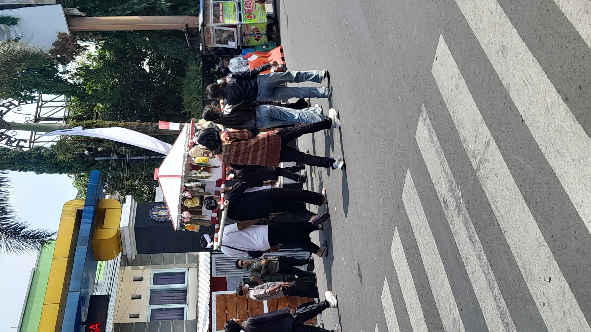 In Sumedang, Indonesia, a group of people are walking across a street, crossing a zebra crossing. In the foreground, several individuals are walking from right to left. The person in the middle is wearing a red and brown patterned long-sleeved shirt and black pants. To their right, a person in a black leather jacket and jeans walks with their back to the camera. To the left of the patterned shirt wearer, a man in a white t-shirt and black pants carries a long white pole over his shoulder. He is accompanied by a man in a black outfit also carrying a pole. A few other people are visible further back, including a person in a black jacket walking away from the camera and a person in a black outfit and a white head covering.
In the background, there is a white and red canopy structure with items hanging from it, possibly for sale. Behind this structure is a building with a blue and yellow facade and a large archway. Further in the background, trees and some street vendors with carts are present. One cart has green and yellow signage that reads "TAKTOYANI" and "TA KOTAKI". The overall environment appears to be a street in an urban setting on a sunny day.