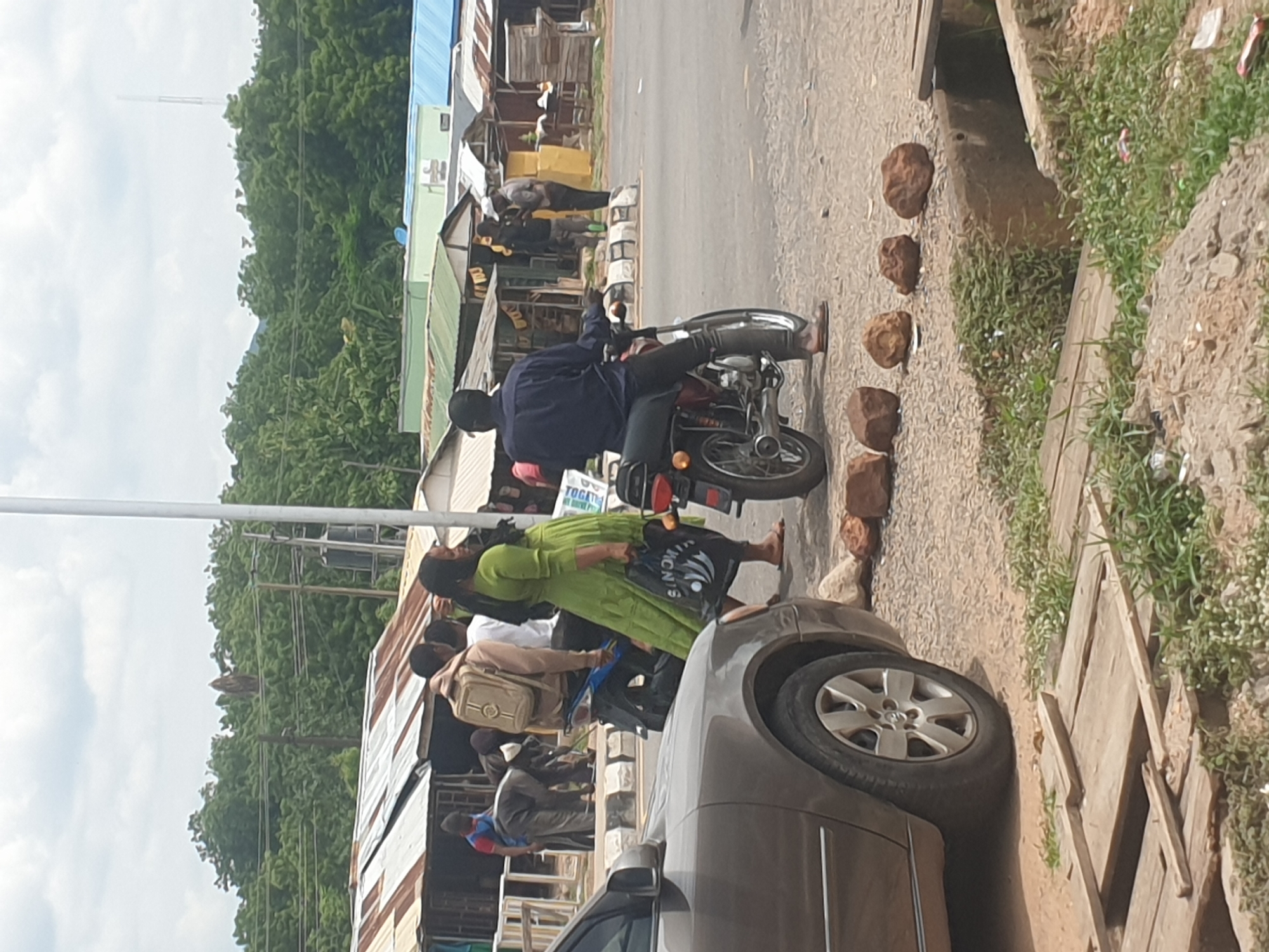 The image shows a street scene in a city with buildings on both sides of the road. A man is sitting on a motorcycle, and several people are standing or walking nearby. A car is partially visible in the foreground. The environment includes trees, utility poles, and a drainage ditch with rocks lining the edge.