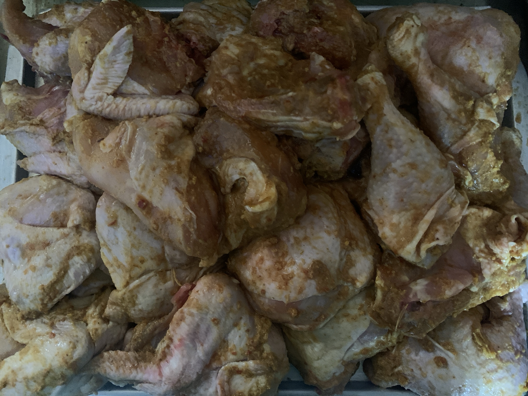 A close-up, overhead shot displays a large quantity of raw chicken pieces, cut into various sections such as wings, drumsticks, and thighs. The chicken is coated in a marinade or spice rub that appears to be a mixture of light brown and yellow hues, with some red flecks, suggesting the presence of turmeric, chili, and other spices. The chicken pieces are densely packed in a metal tray or container, filling the entire frame. The lighting is even, casting minimal shadows, and the focus is sharp, highlighting the texture of the raw meat and the seasoning. The scene is devoid of any human presence or specific environmental details that would indicate a location within the city of Fajo 2 Division Range, Nigeria, or any other specific geographical context.