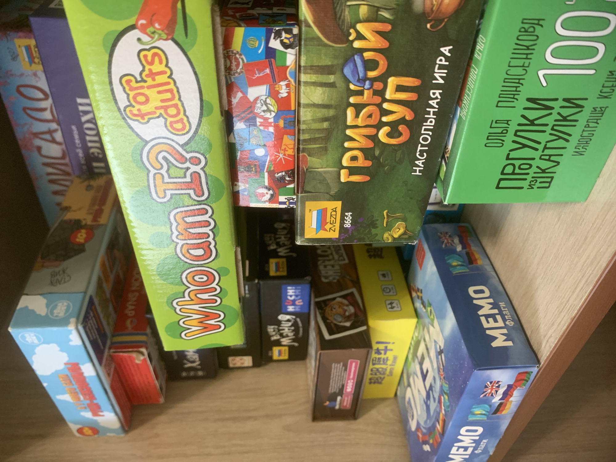 The image shows a shelf filled with board games. The games are stacked tightly together, with only the spines visible. The titles of some of the games include "Alisalo", "Jon Snap", "Who am I? for adults", "Mania Muchi!", "Sherlock Holmes", "Gribnoy Sup" ("Mushroom Soup" in Russian), "Progulki iz shkatulki 100" (possibly referring to the number 100), and "Memo Flags." The games appear to be a mix of Russian and English titles. The shelf is made of wood. Given the Russian titles of some of the games, this image was most likely taken in Russia.