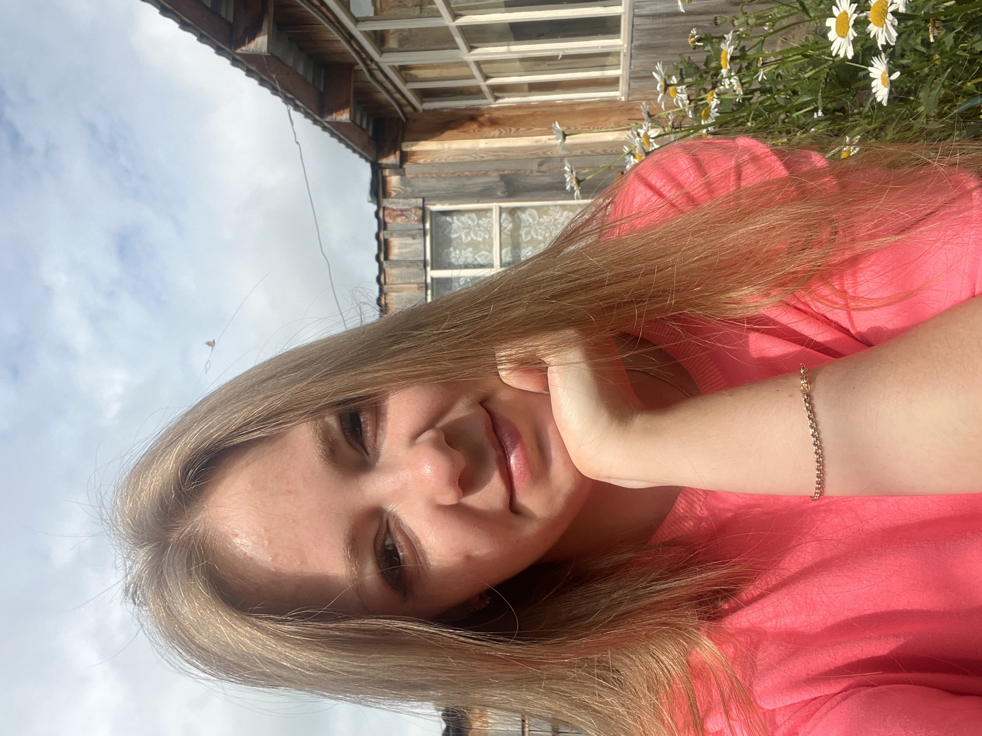 A young blonde woman with fair skin is pictured from the chest up, taking a selfie outdoors. She is wearing a bright pink crewneck shirt and a delicate gold bracelet on her right wrist. Her left hand rests on her chin, and she is smiling slightly. Her long blonde hair falls around her shoulders. The background suggests a rural or suburban setting, with a rustic wooden building and a patch of white daisies. The sky is partly cloudy. The image appears to be taken on a sunny day. The context of Kovriga, Russia, implies this is likely a personal photograph taken at a home or in a nearby outdoor space.
