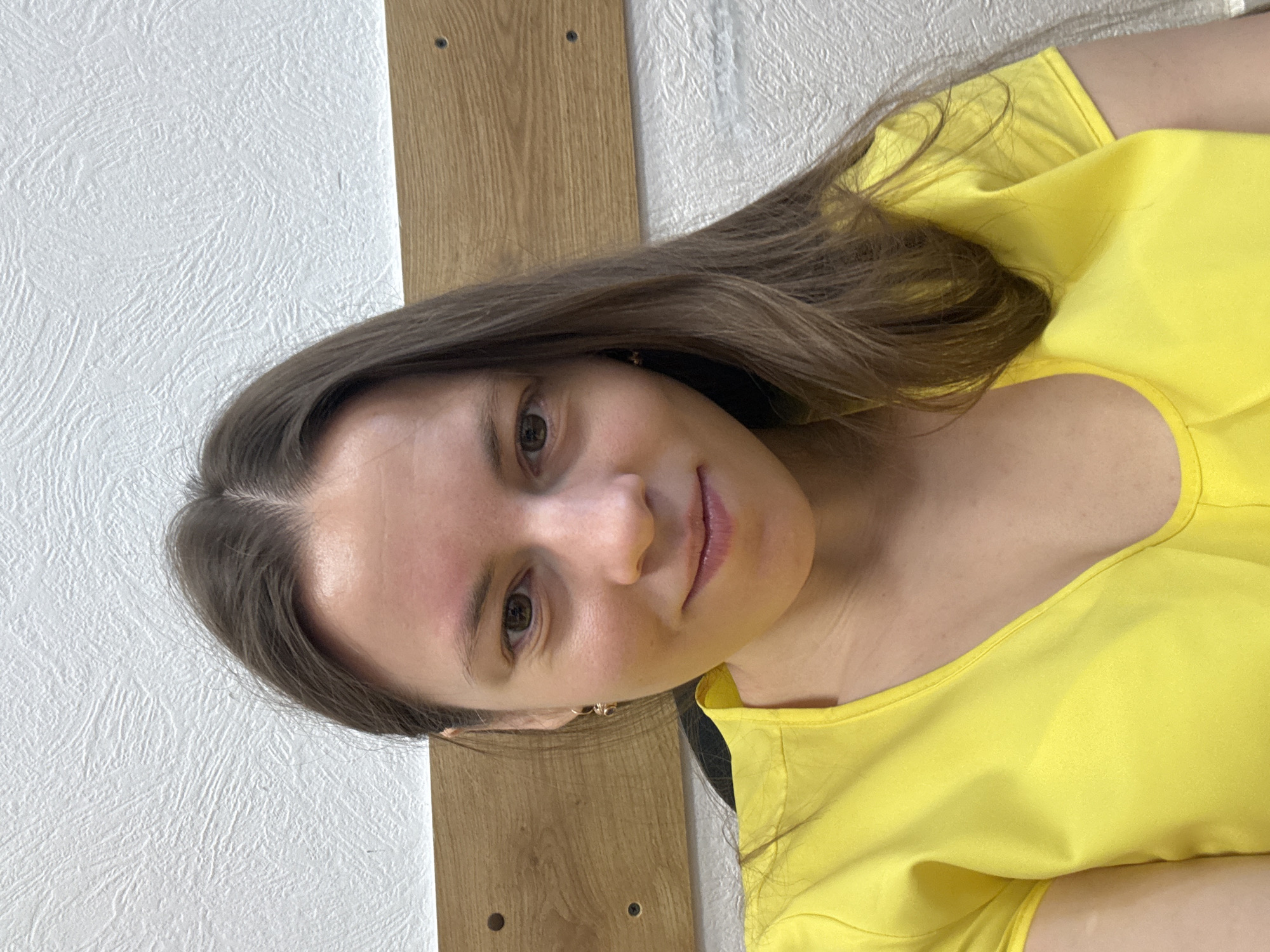 A woman is seen in a close-up shot, wearing a yellow top. Her brown hair is styled loosely around her face. The background includes a white textured wall and a section of wooden board with visible screw holes. The image does not provide any information regarding the location in Chaikovsky, Russia.