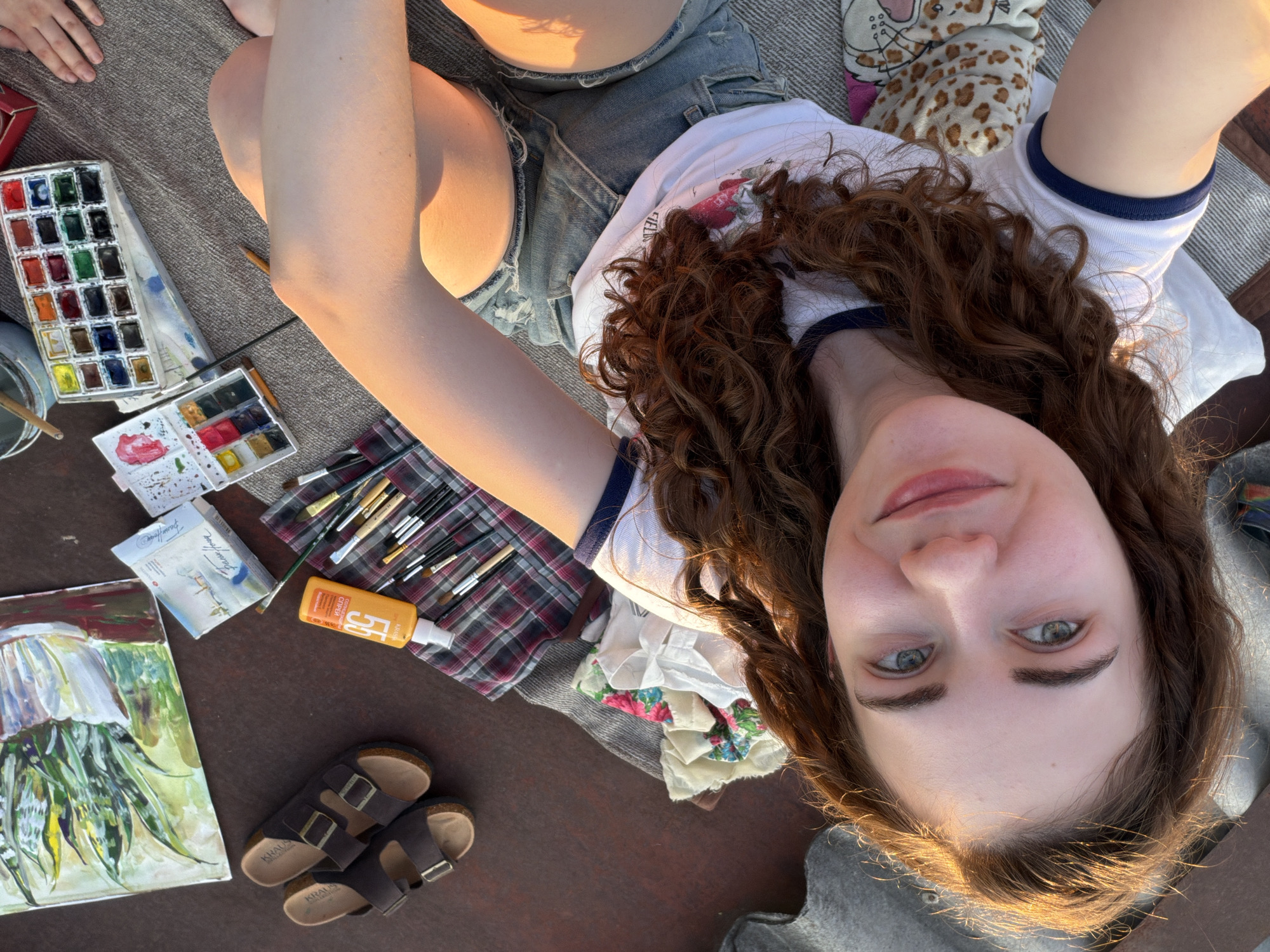 The image depicts a young woman with curly brown hair and blue eyes, wearing a white t-shirt with a blue trim and denim shorts. She is sitting on a blanket outdoors, with a pillow and some patterned fabric nearby. Various art supplies are spread around her, including paintbrushes, a bottle of sunscreen, a watercolor paint set, and a partially completed painting of a plant in a pot. A pair of brown sandals is also visible. The lighting suggests it is either sunrise or sunset, casting a warm glow on the scene. The location is not specified within the image or any provided context.