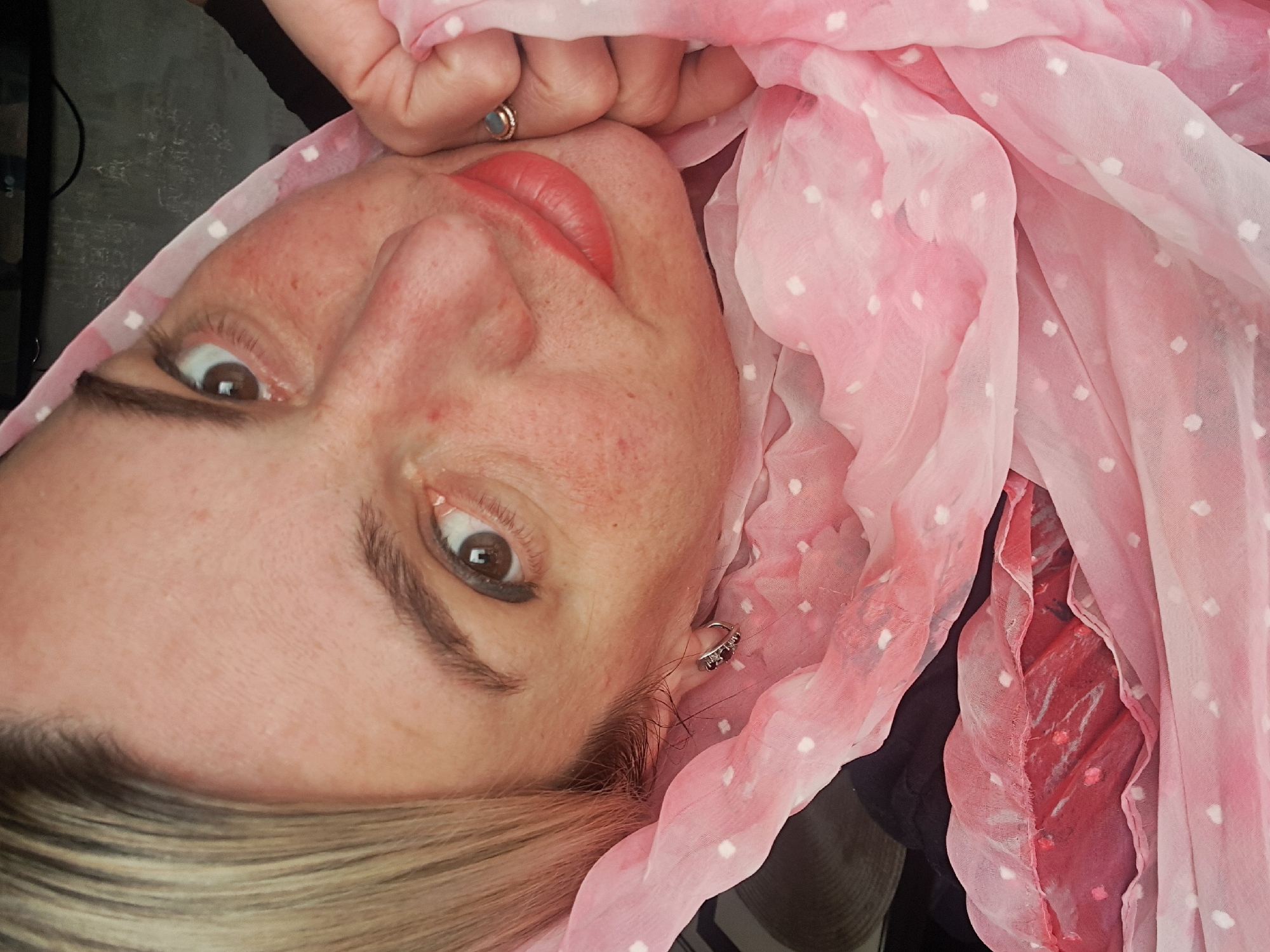 A woman's face is the focal point of the image, oriented at a diagonal. She has fair skin, brown eyes, and blonde hair. Her lips are painted a shade of red. She is wearing a ring on one of her fingers. A light pink scarf with white polka dots is draped around her head and neck. An earring is visible on one ear. The background is indistinct, but appears to include dark and red objects, and a black surface. There is no indication of a location, especially not Elektrostal, Russia.